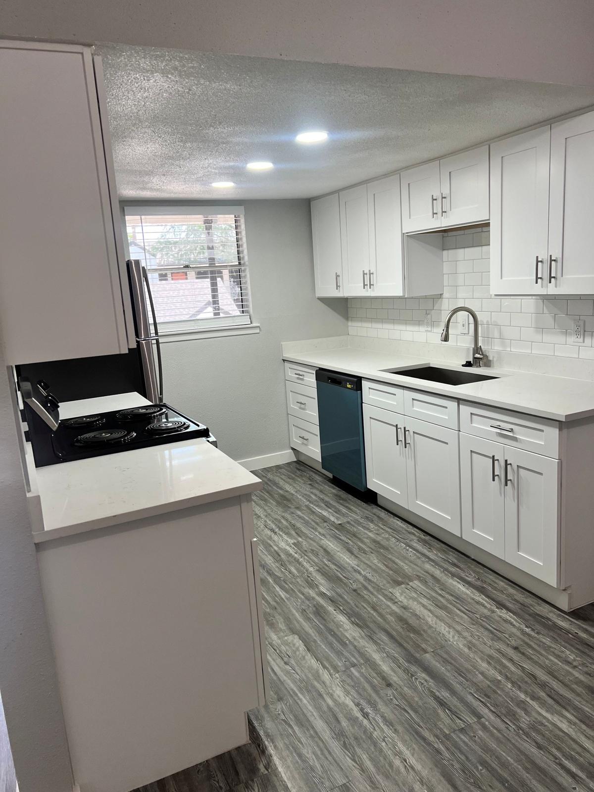 Modern kitchen featuring white cabinetry, a gray and white tiled backsplash, and stainless steel appliances. Natural light enters through a window, illuminating the space. The floor is covered with gray vinyl planks, and there's a countertop on one side with a stove.