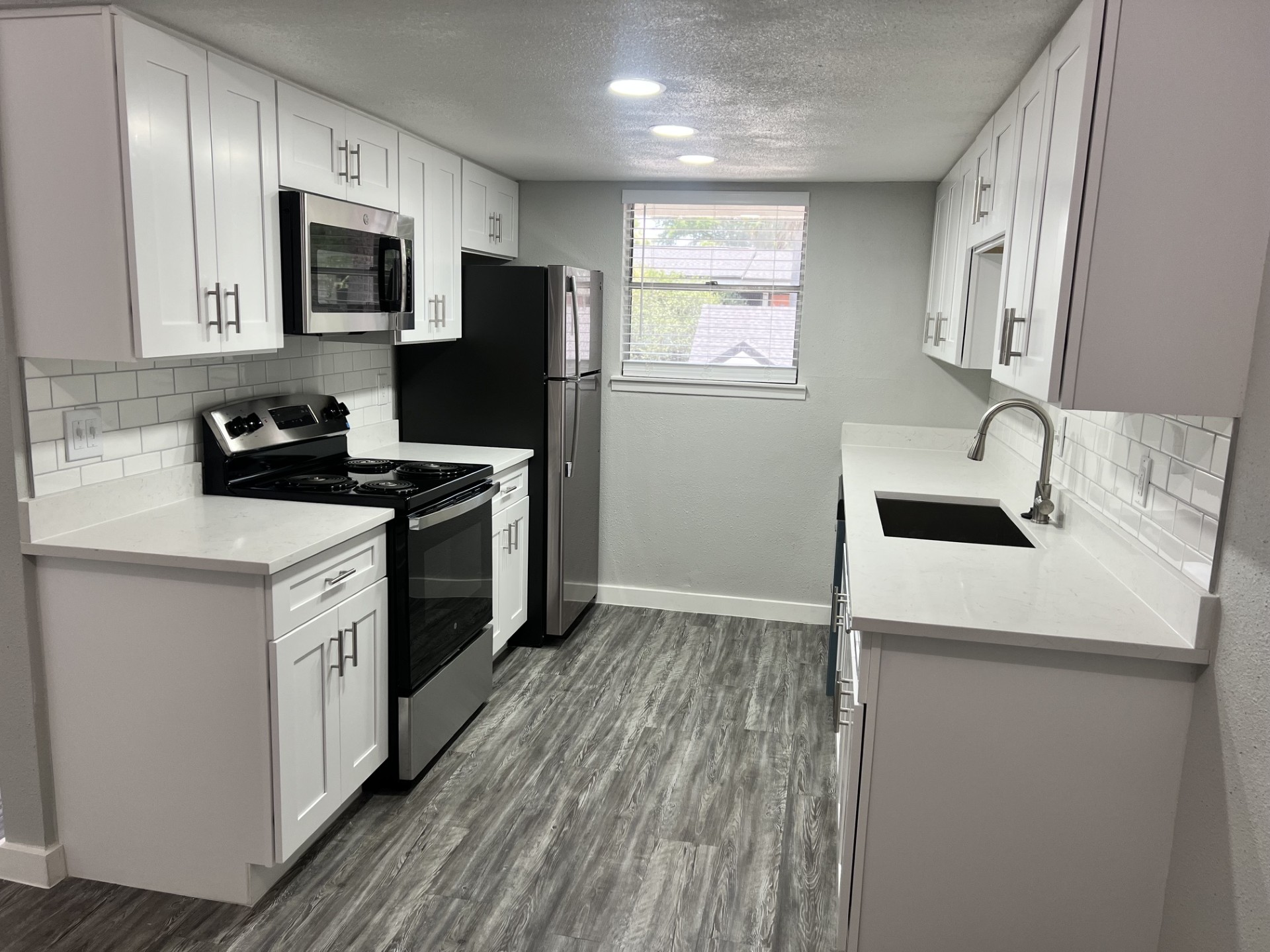 Modern kitchen featuring white cabinetry, stainless steel appliances including a microwave, oven, and refrigerator. The countertops are a light-colored stone, and a single basin sink is integrated into the counter. The floor is a dark wood-like laminate, and there is a window allowing natural light to enter.