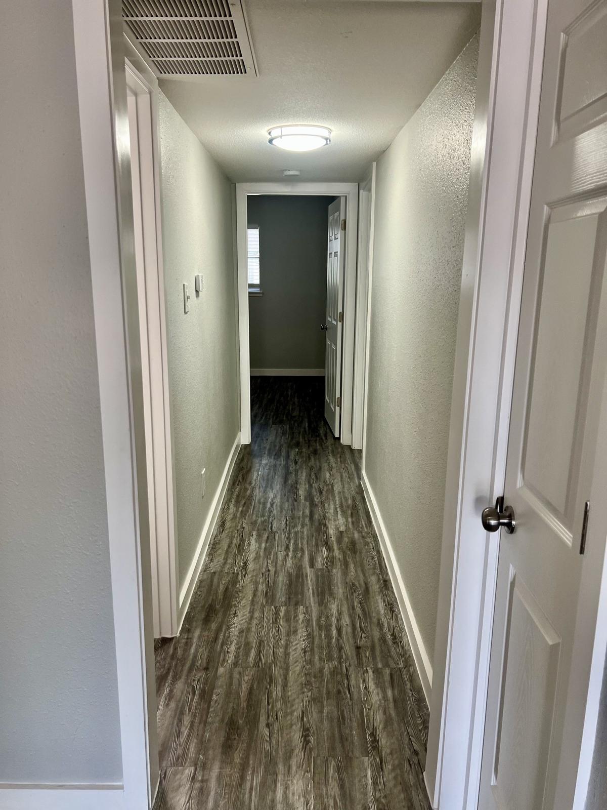 A well-lit hallway with light gray walls and a dark wood-like floor. The corridor features a ceiling light, doors on both sides, and a window at the far end, creating an open and inviting atmosphere.