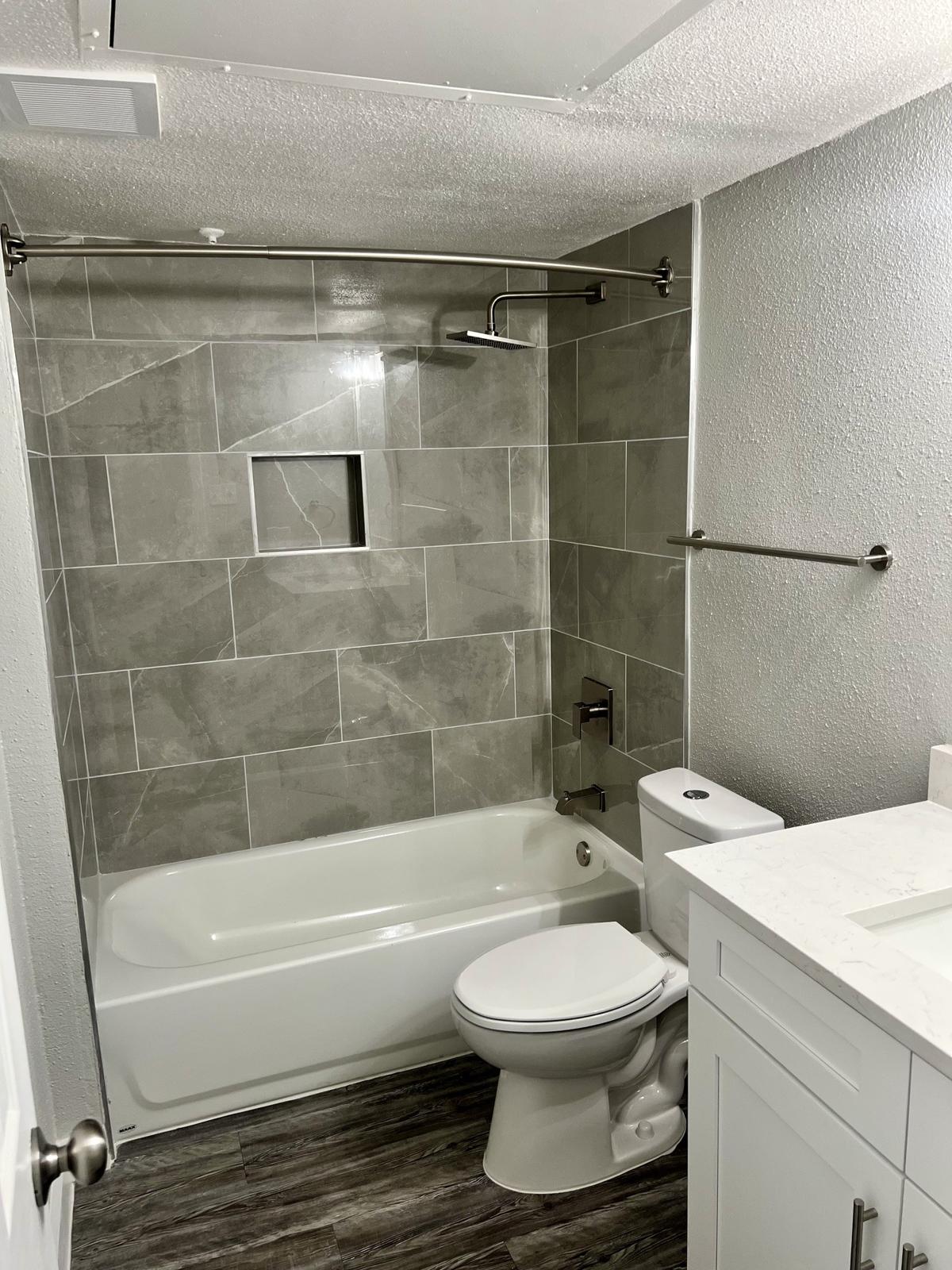 A modern bathroom featuring a bathtub with a shower head, gray tiled walls with a built-in shelf, a toilet, and a white vanity with a sink. The flooring is wood-like, and the overall design is clean and contemporary. The walls have a smooth finish, and there is adequate lighting.