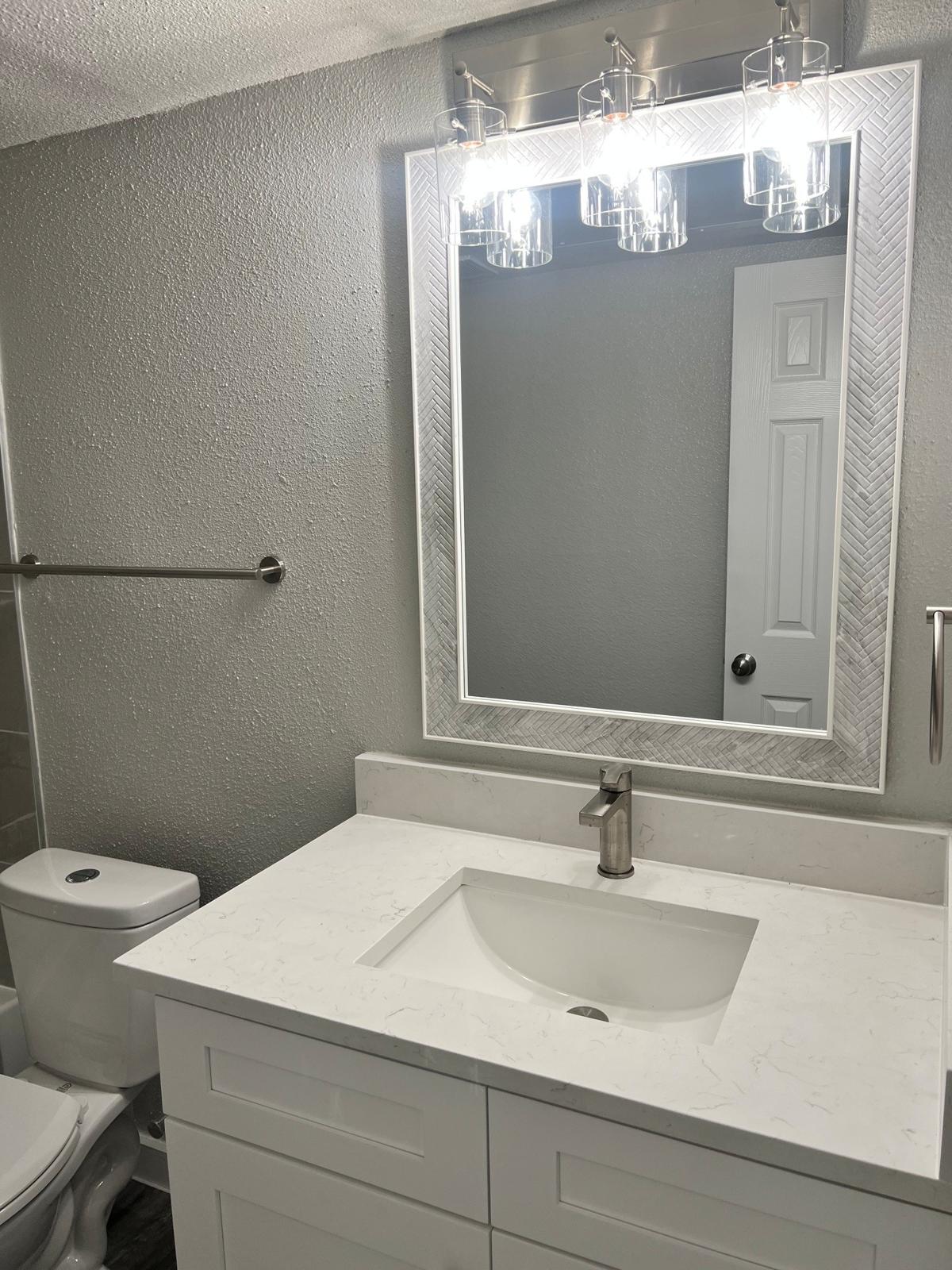 A modern bathroom featuring a white vanity with a sink, a large mirror surrounded by a decorative frame, and three pendant lights above. A toilet is visible in the background, along with a light gray wall and tiled flooring. The overall look is clean and contemporary.