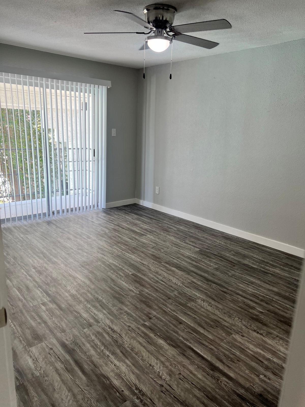 A spacious, empty room with gray walls and a ceiling fan. The floor features wood-like laminate, and there are sliding glass doors with vertical blinds leading to an outdoor area. Natural light is visible through the doors, creating a bright and airy atmosphere.