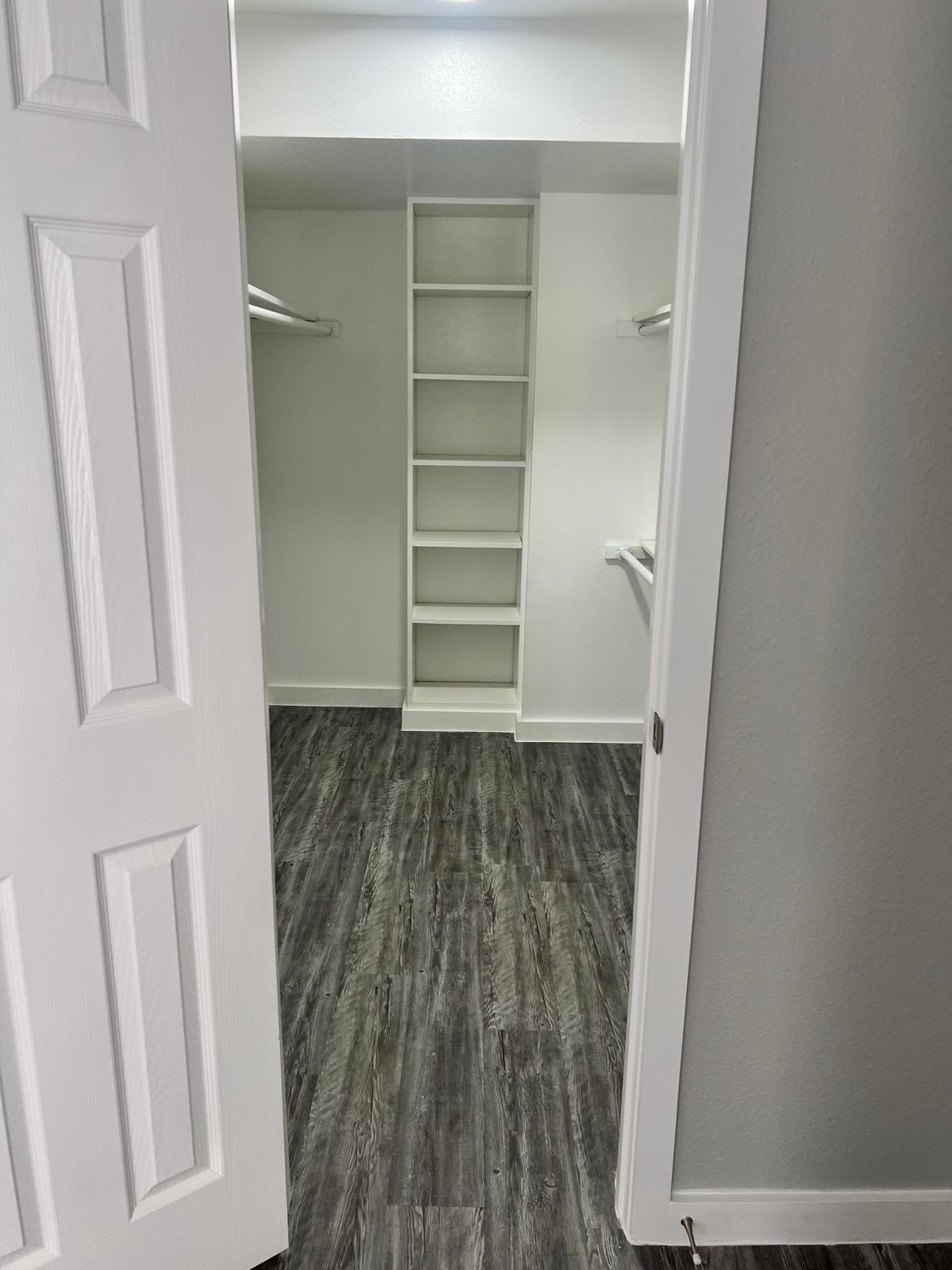 A spacious walk-in closet with white walls and shelves. The closet features wooden flooring and is well-lit from overhead. Two doors are open, revealing organized shelving in the center and hanging rods on either side, creating ample storage space.