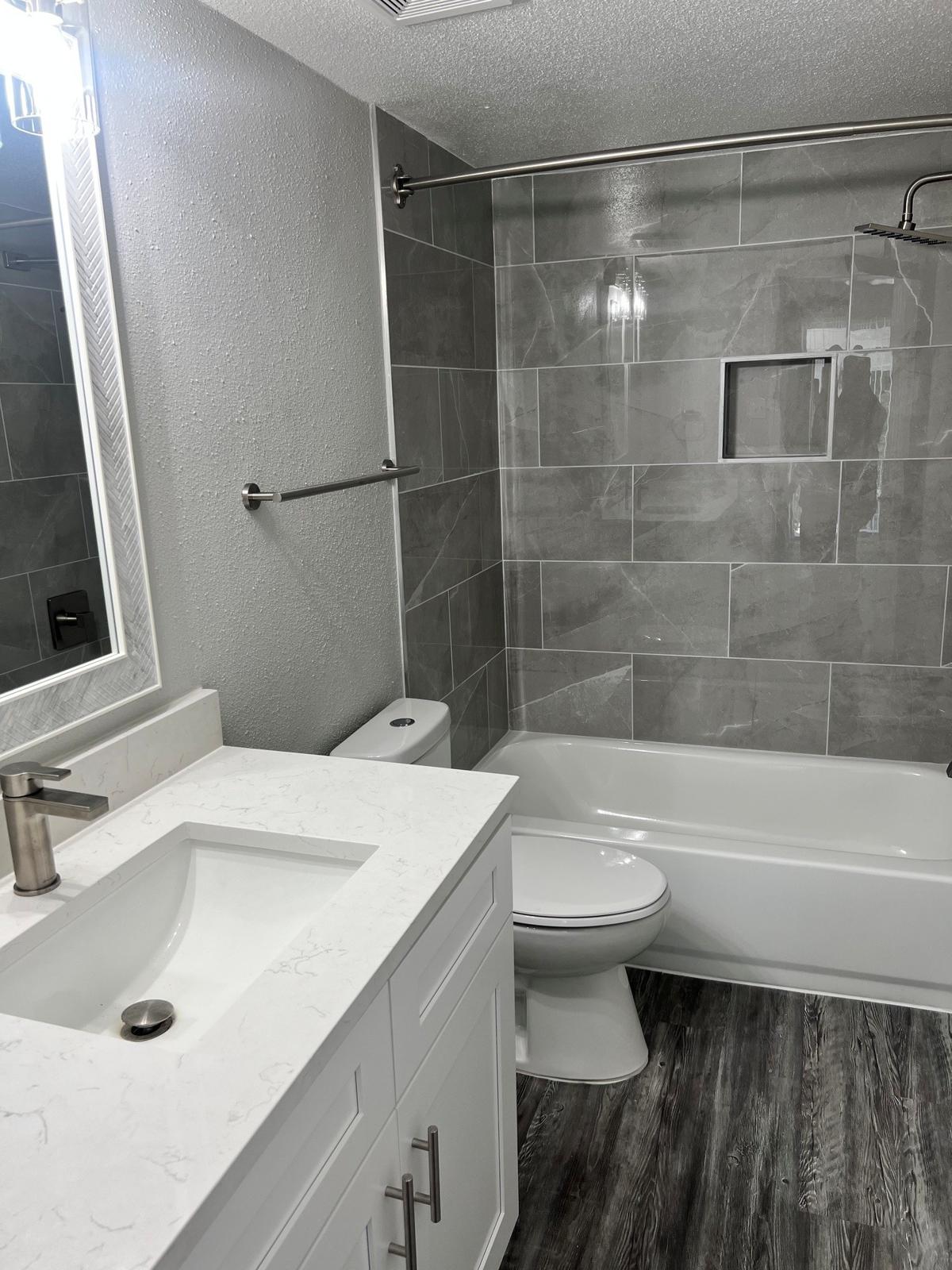 A modern bathroom featuring a sleek vanity with a marble countertop and a single basin sink. The bathtub is set against a backdrop of gray tiled walls, with a glass shower door. A white toilet is positioned nearby, and the floor has a dark wood-like appearance. A well-lit mirror hangs above the sink.