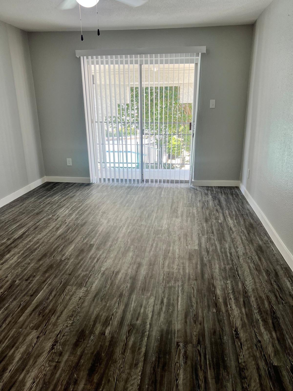 A spacious, empty room with light gray walls and dark wood-like flooring. A ceiling fan is visible, and large sliding glass doors with vertical blinds open to a patio area overlooking a pool. Natural light enters the room, enhancing its bright and airy atmosphere.