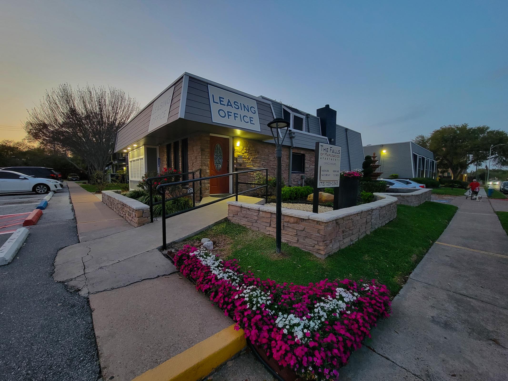 Leasing office of an apartment complex, featuring a well-maintained garden with colorful flowers in front. The building has a sign indicating its purpose and is set against a twilight sky. Pathways lead up to the entrance, with nearby parking and residential units visible.