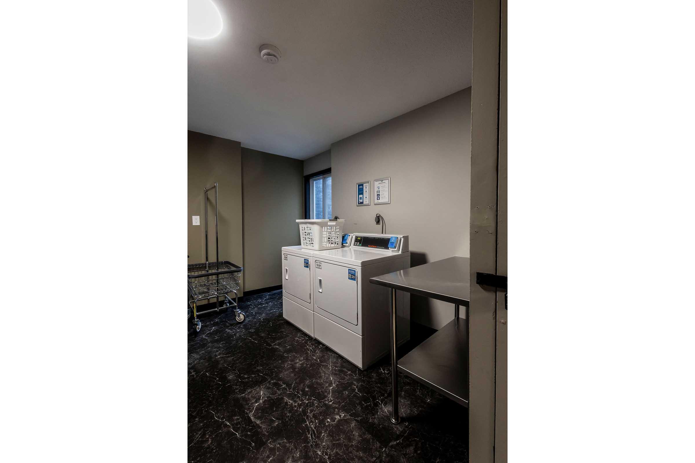 A laundry room featuring a washing machine, dryer, and a metal table. There is a laundry cart nearby, and a window allows natural light into the space. The walls are painted in a neutral color, and the flooring is dark.
