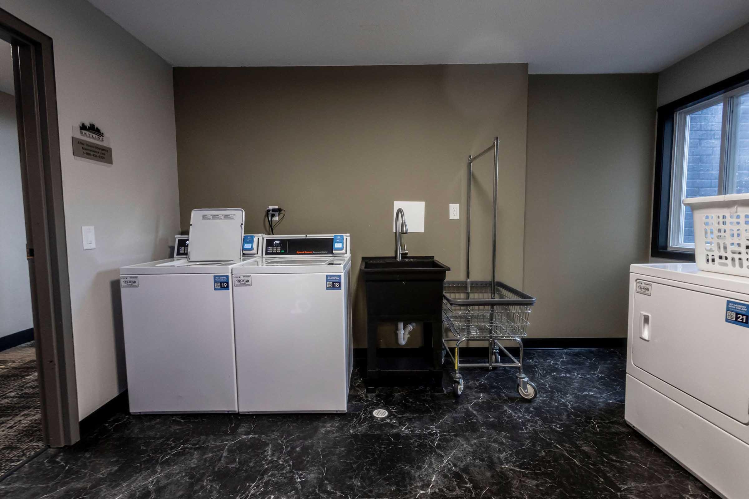 Laundry room featuring two white washer and dryer units, a black utility sink, a metal cart, and a laundry basket. The room has dark marble flooring and neutral-colored walls, with a window allowing natural light to enter.
