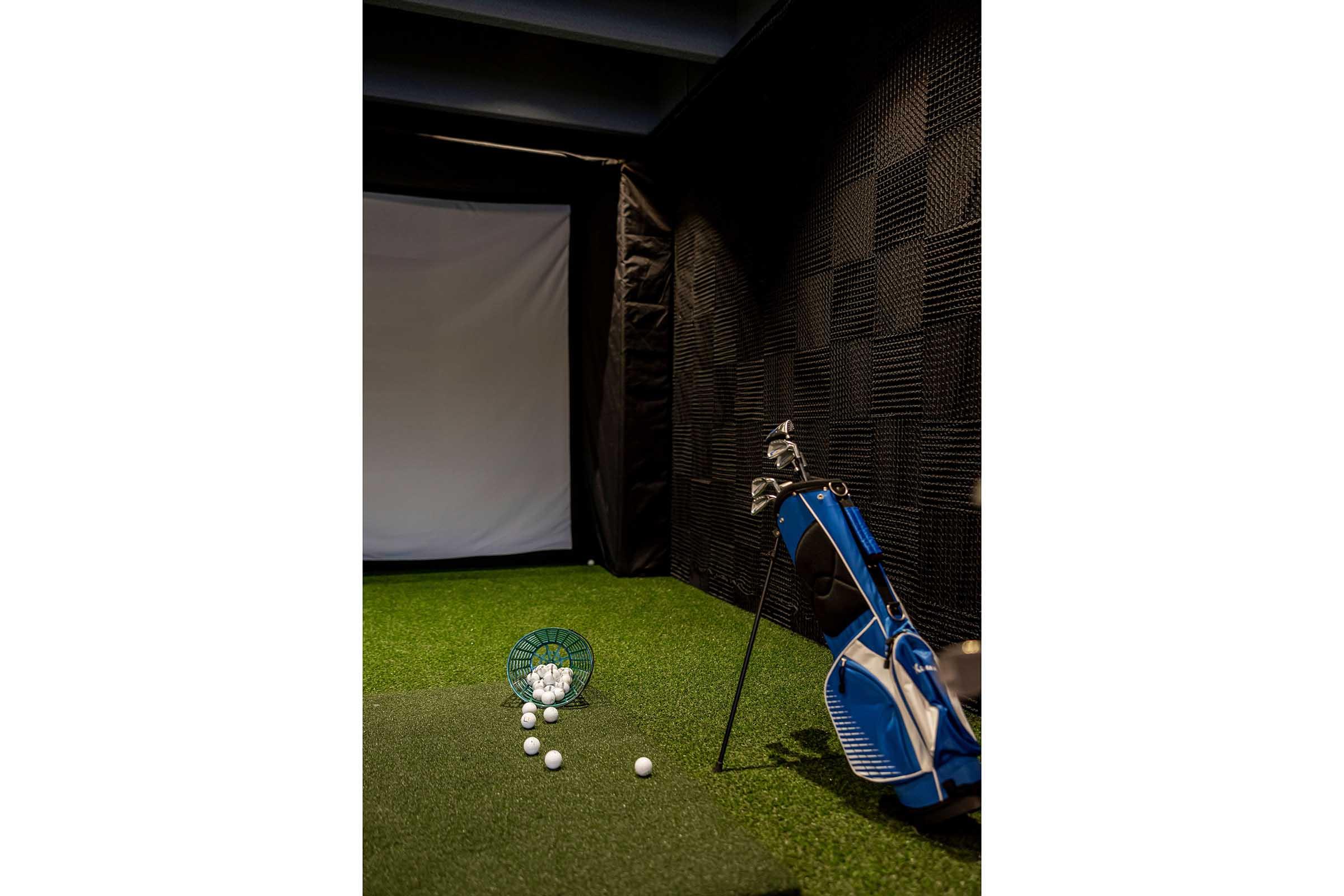 Indoor golf setup featuring a hitting area with artificial grass, a golf bag with clubs, and a basket of golf balls. The background includes a large screen for simulation, with soundproofing material on the walls. Lighting is soft to create a relaxed atmosphere for practice.