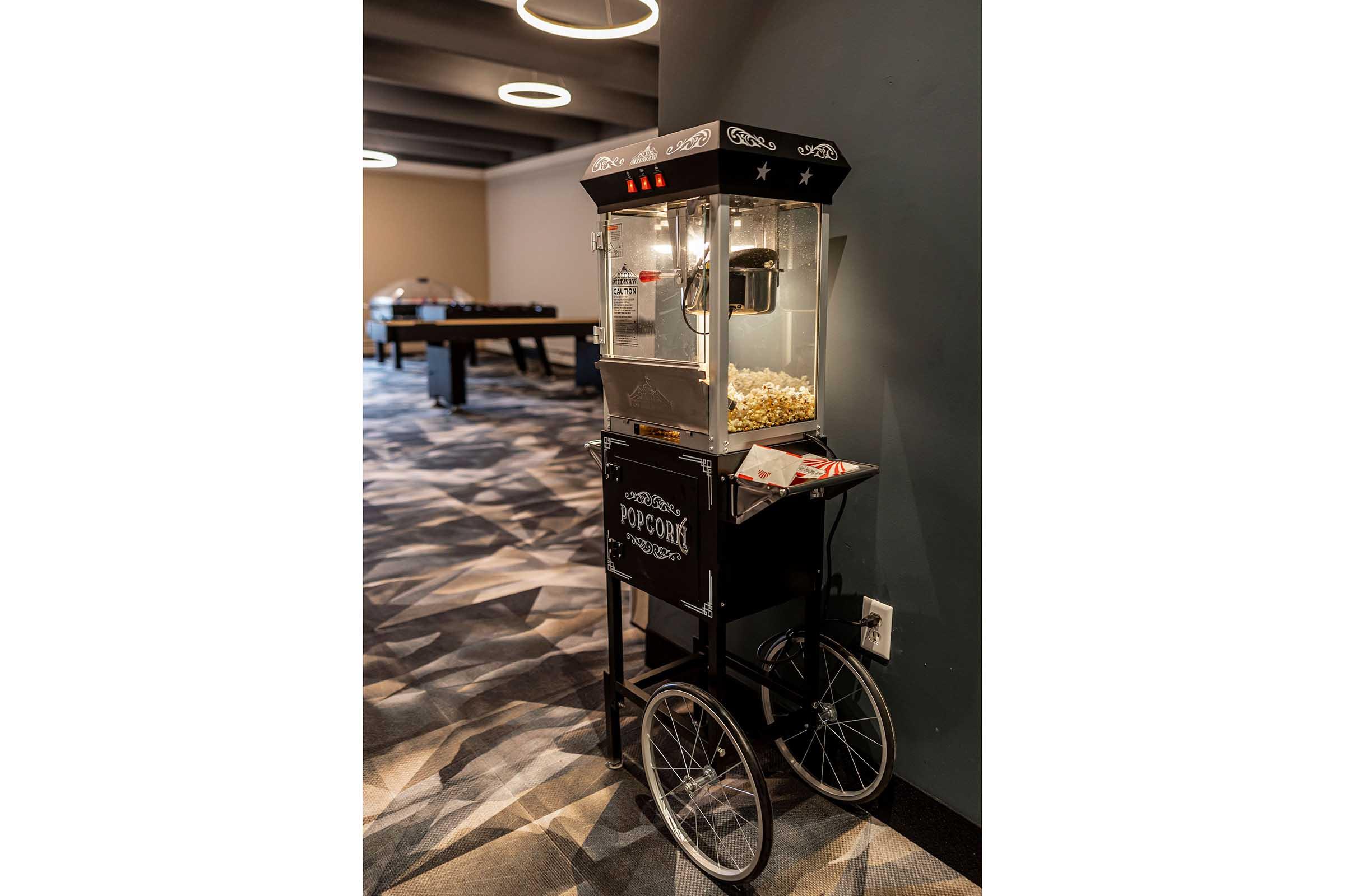 A vintage-style popcorn machine on a wheeled cart, with a glass top showcasing freshly popped popcorn. The cart is black with decorative elements, and there are bags of popcorn nearby. The setting features modern decor with soft lighting and a patterned carpet.