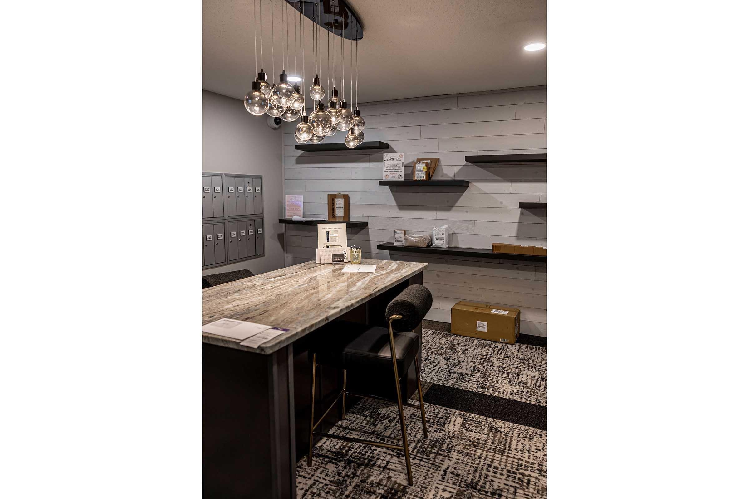 Interior of a modern reception area featuring a granite countertop with a sleek black chair, decorative pendant lights hanging from the ceiling, and black shelving units against a light-colored wall. There are organized mailboxes and packages displayed on the counter, creating a welcoming atmosphere.