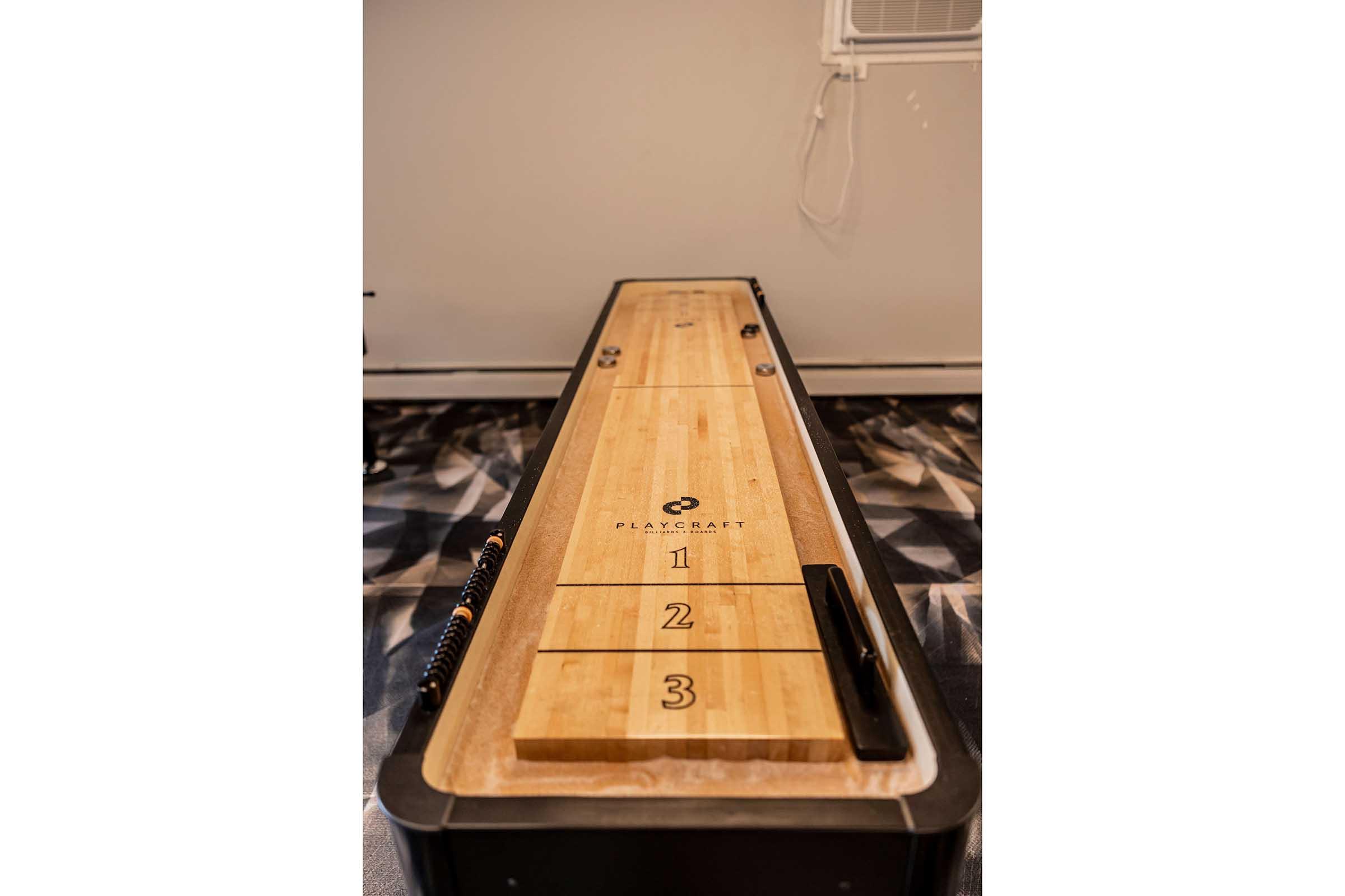 A wooden shuffleboard table with three numbered scoring sections. The table has a smooth surface and is positioned in a room with patterned flooring. An air conditioning unit can be seen in the background, and the table features the brand name "Playcraft" prominently displayed.