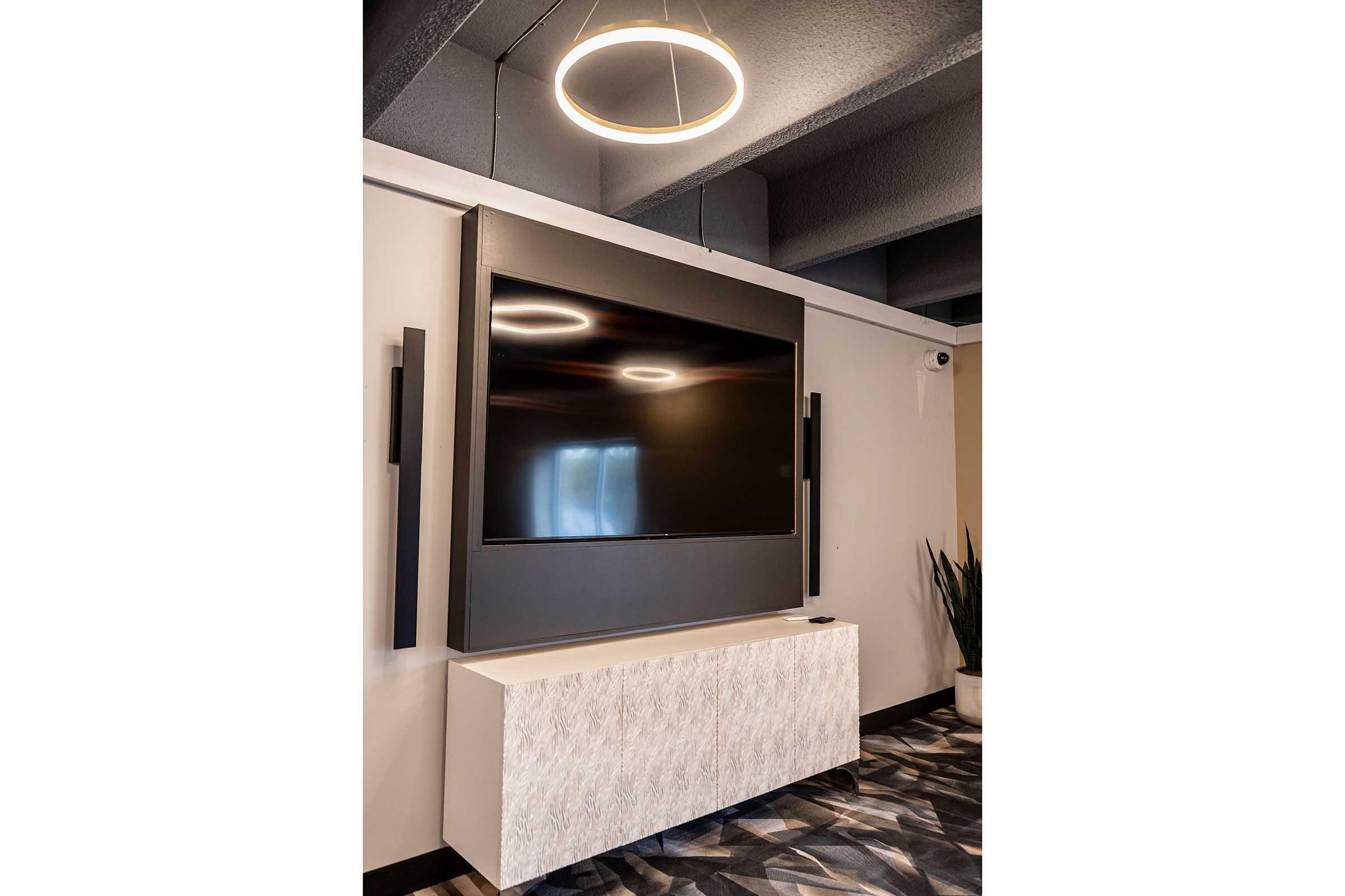 A modern television mounted on a wall, with a sleek black casing and a white decorative cabinet below. The space is well-lit with a circular pendant light overhead, and a plant is visible in the corner. The flooring features a contemporary patterned design.
