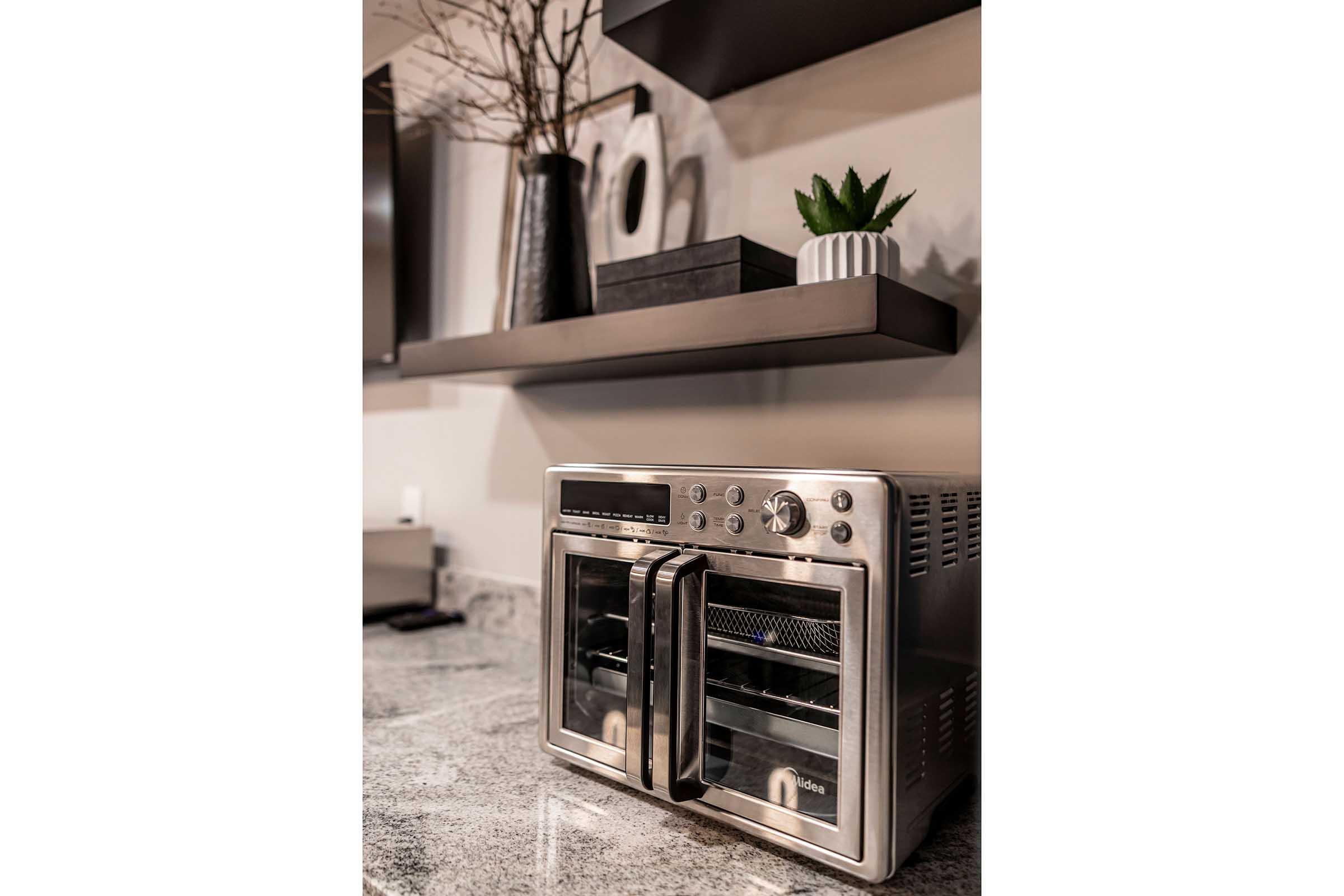 A modern kitchen countertop featuring a silver toaster oven with dual doors, placed on a granite surface. Above the toaster oven, there are dark shelves with decorative items, including a small potted plant and a black storage box, adding a sleek and contemporary touch to the kitchen decor.