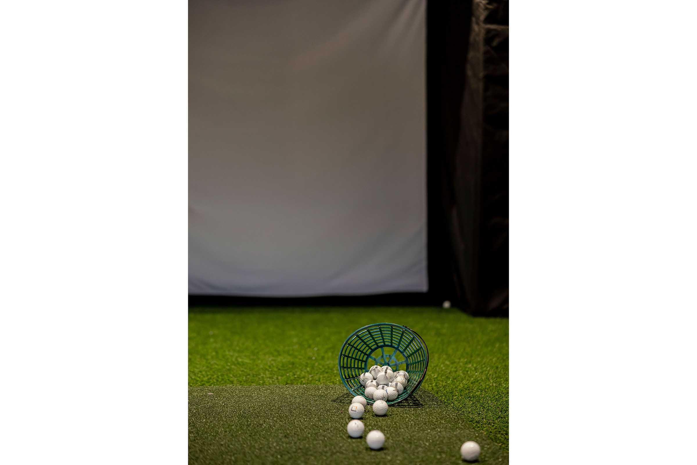 A basket tipped over, scattering white golf balls on a green turf surface, with a blank screen in the background, suggesting a golf practice or simulation area.