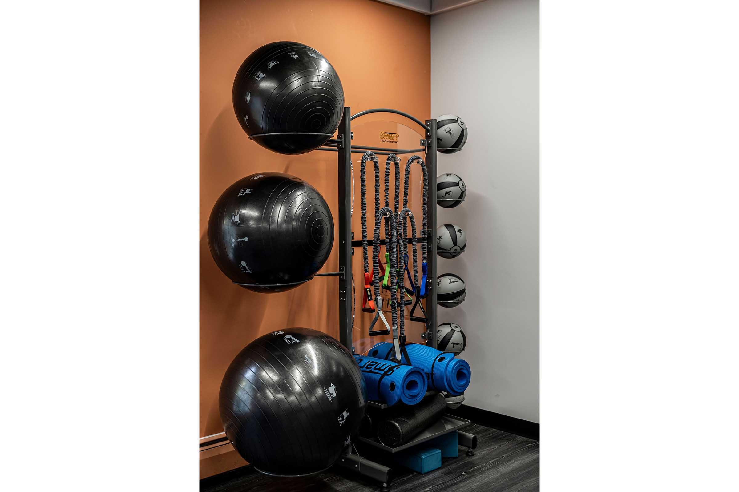 A fitness area featuring exercise equipment, including large black stability balls mounted on a rack, a collection of helmets hanging on a wall-mounted holder, and exercise mats stacked on a shelf, all set against an orange wall.