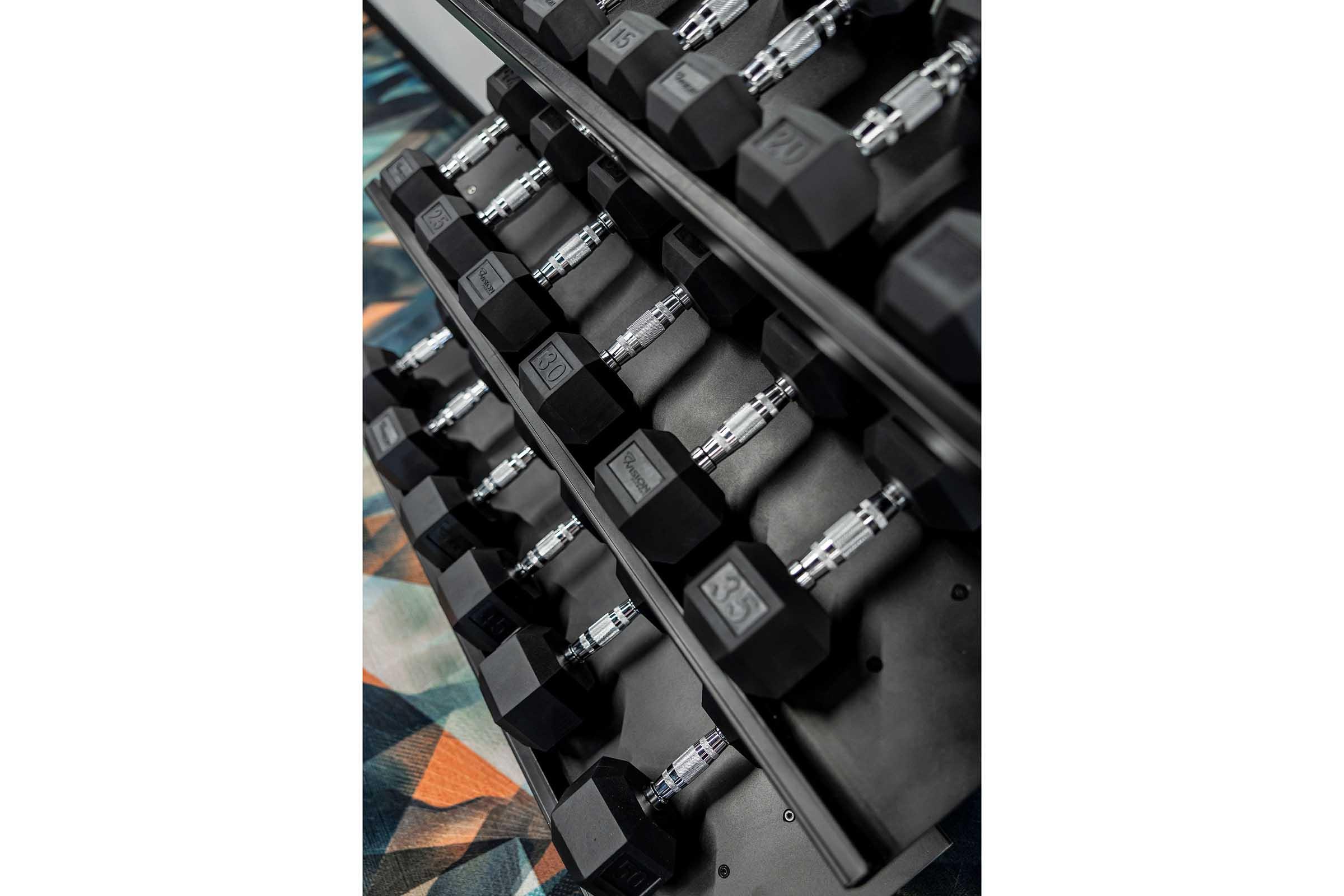 A set of black hex dumbbells arranged neatly on a rack, with a patterned gym mat visible underneath. The weights are labeled with their respective values, providing an organized view of exercise equipment.