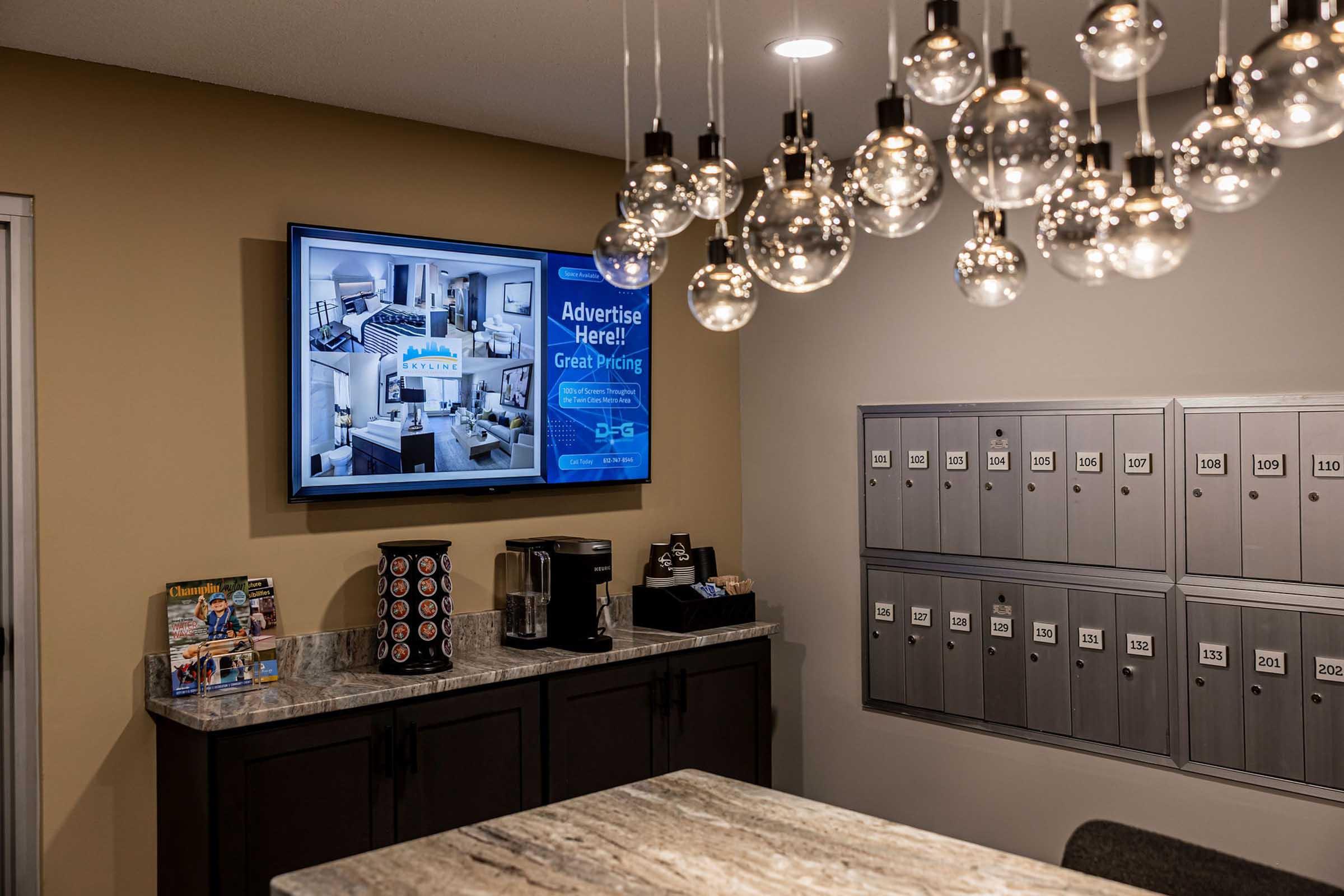 A modern lobby area featuring a television displaying promotional content, a coffee station, and a mail slot wall with multiple numbered mailboxes. The space has stylish pendant lighting and a contemporary design.