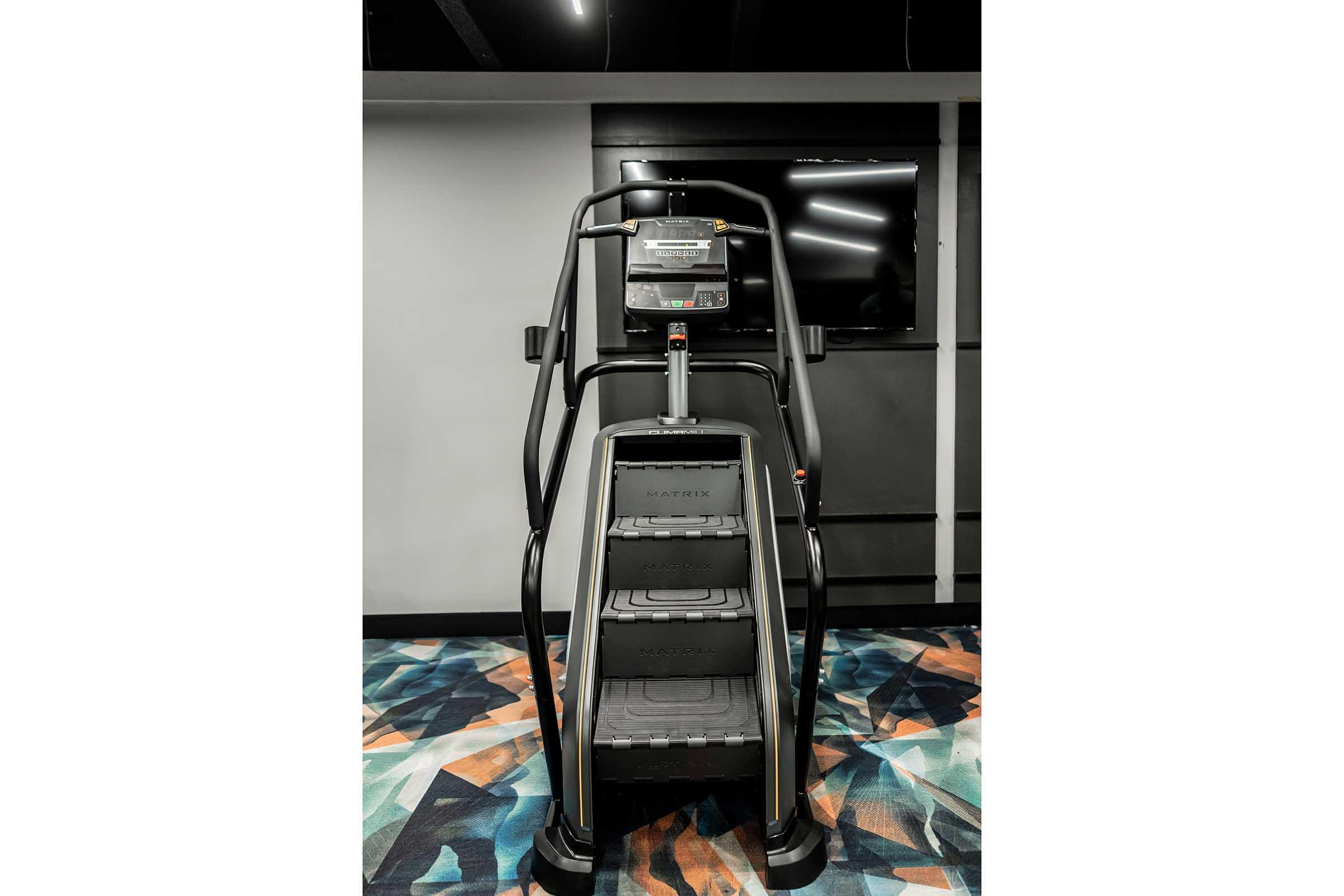 A Matrix stair stepper machine in a modern gym setting, featuring a display screen and a backdrop of dark walls. The floor has a colorful geometric pattern.