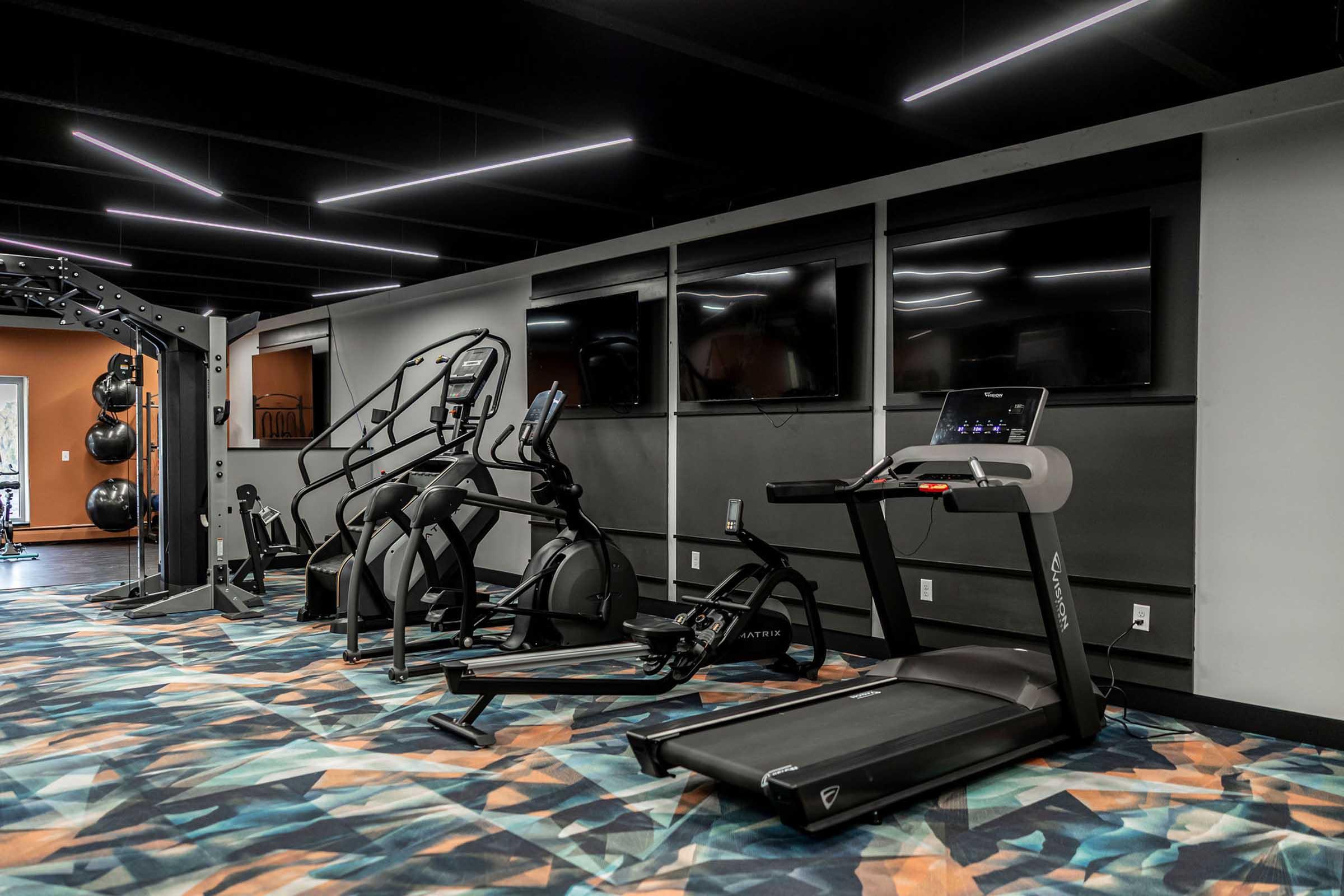 A modern gym interior featuring various exercise equipment, including a treadmill, elliptical machine, and strength training station. The gym has a colorful floor design and is equipped with multiple wall-mounted televisions, enhancing the exercise environment. Bright overhead lighting illuminates the space.