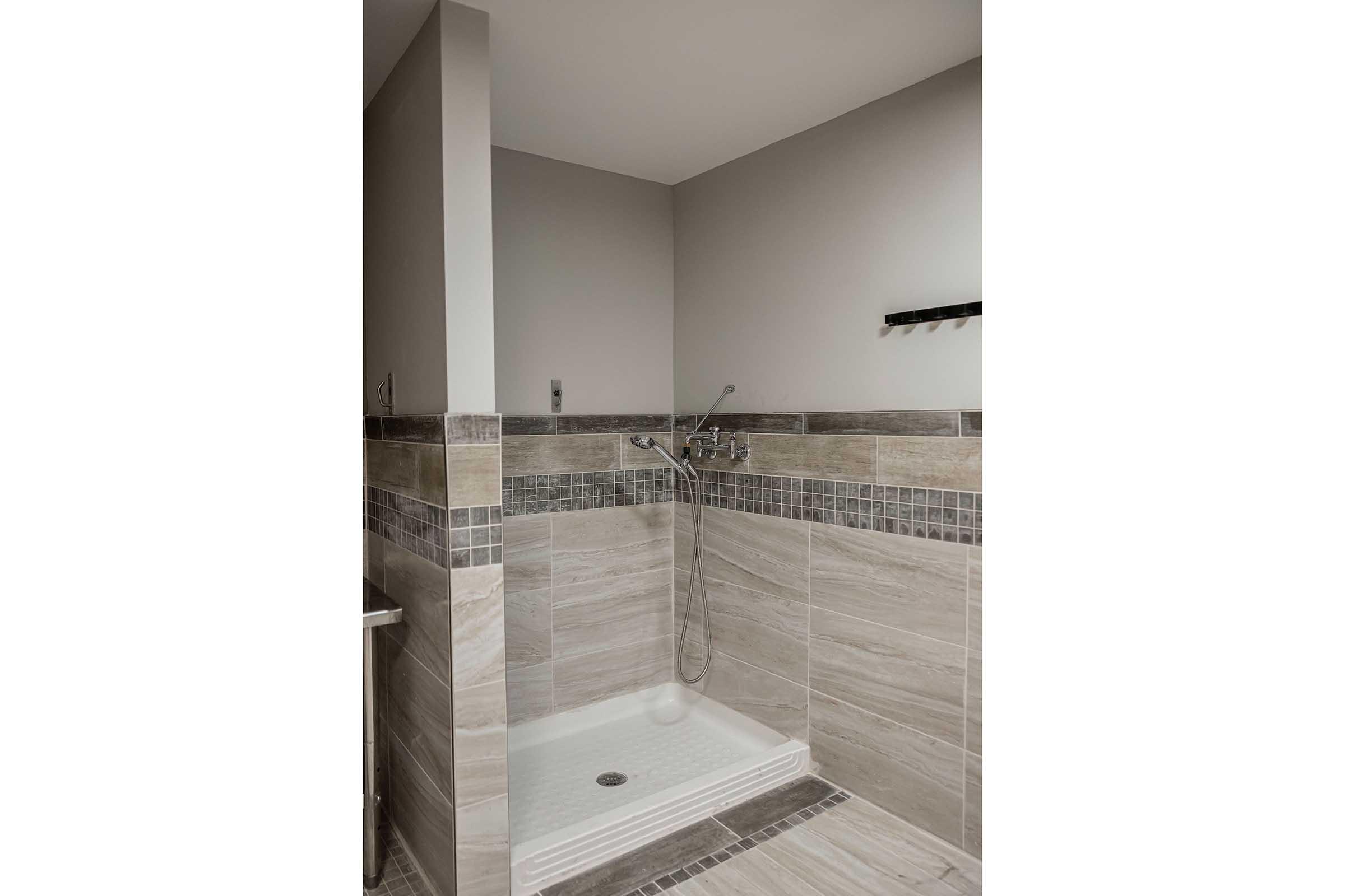 Modern shower stall featuring a tiled interior with gray and beige tones. The shower includes a handheld showerhead, a wall-mounted faucet, and a sleek design with a glass panel. There is a small shelf mounted on the wall for toiletries. The overall aesthetic is contemporary and minimalistic.