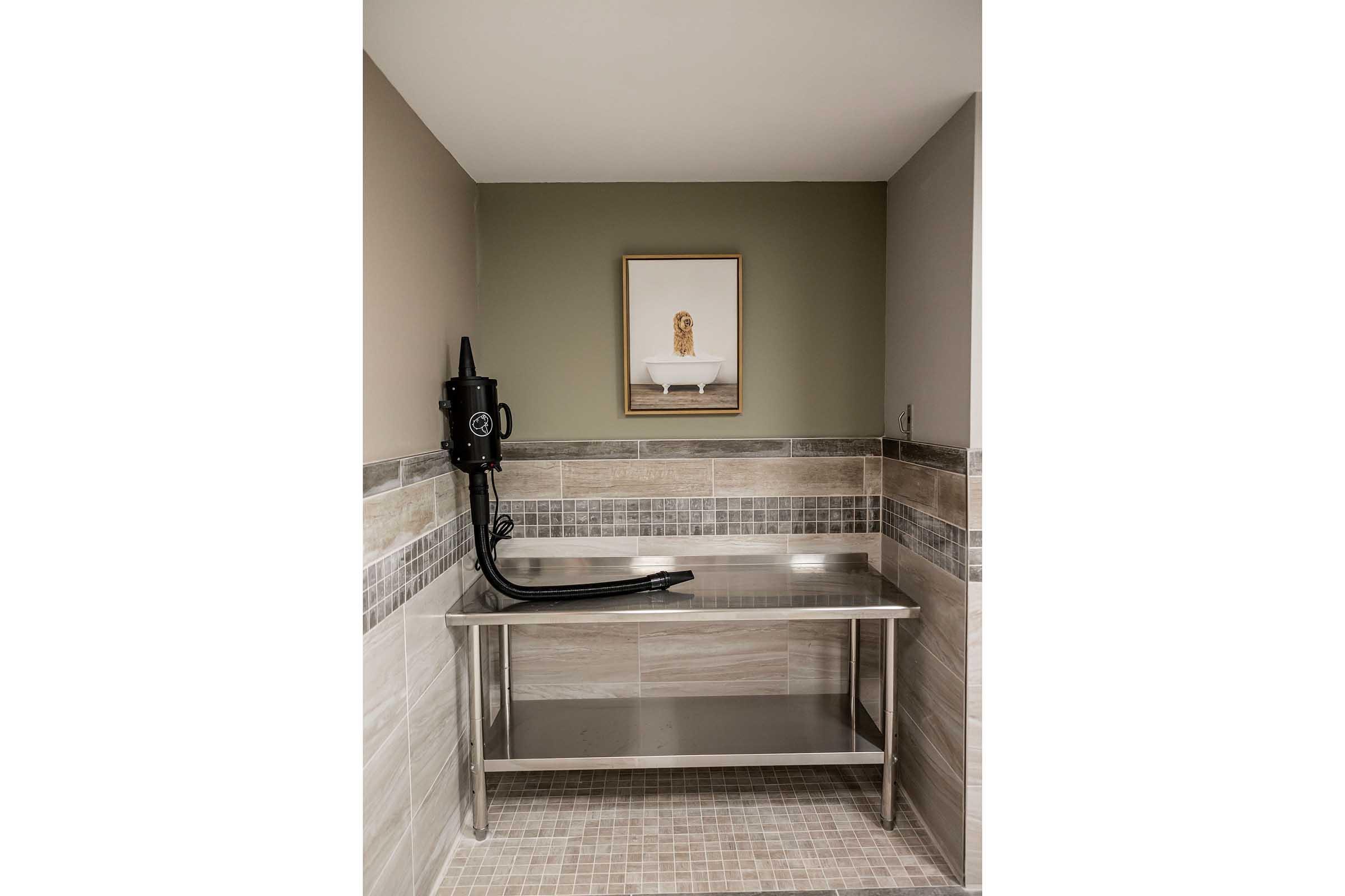 A modern bathroom setting featuring a stainless steel countertop with a black hand dryer mounted on the wall. The walls are adorned with neutral-colored tiles and a framed artwork depicting a bathtub, adding an elegant touch to the space.