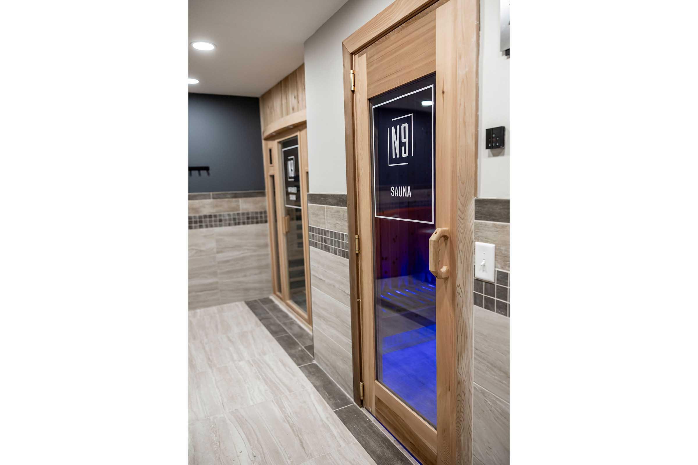 Two wooden sauna doors with glass panels, labeled "SAUNA," situated in a modern hallway. The walls feature a mix of light tiles and dark accents, while subtle lighting creates a calming ambiance.