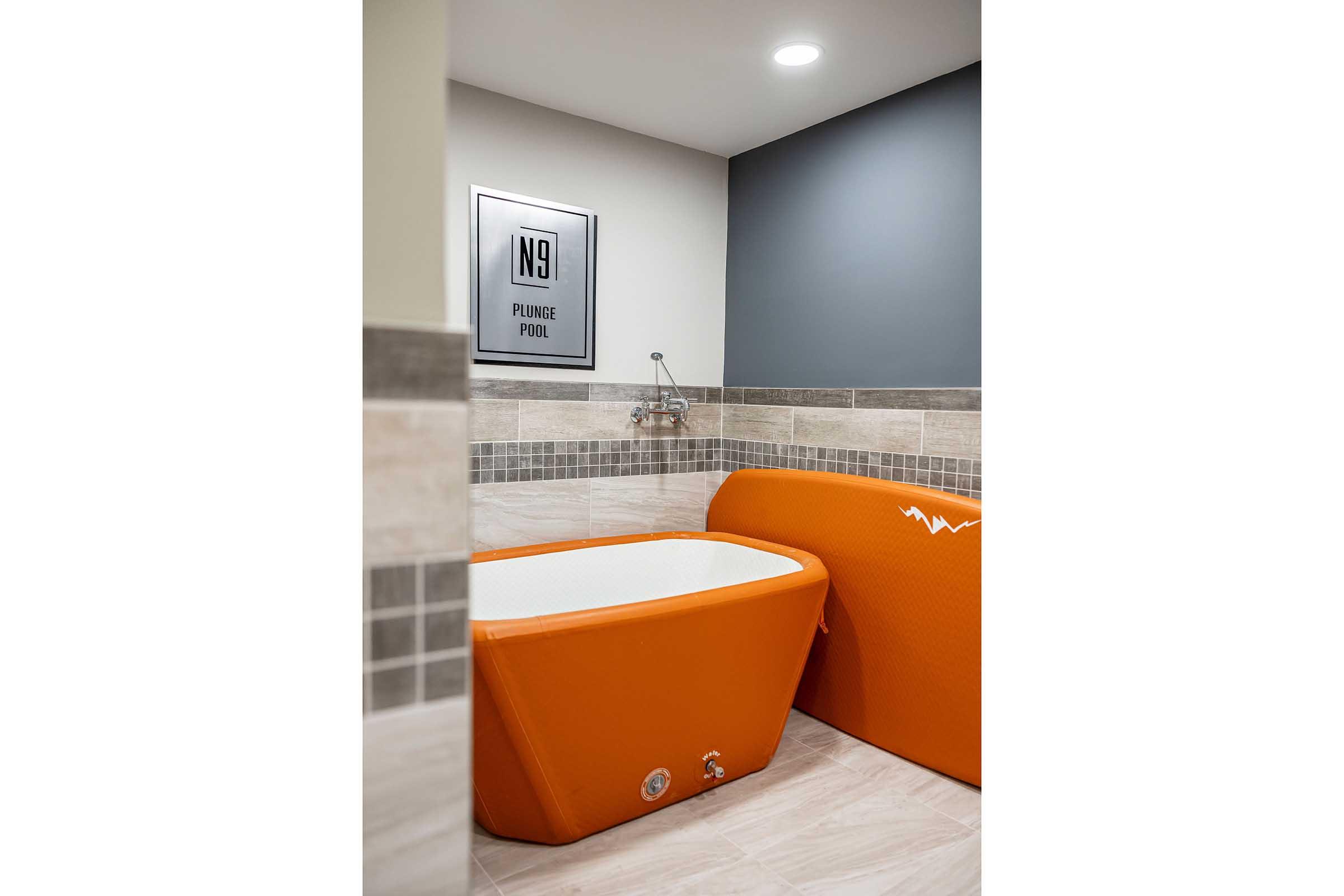 A modern bathroom featuring a stylish orange plunge pool, white interior, and tiled walls. A framed sign labeled "N9" is mounted on the wall, adding a contemporary touch to the sleek design. The space is well-lit with a ceiling light, creating a bright and inviting atmosphere.