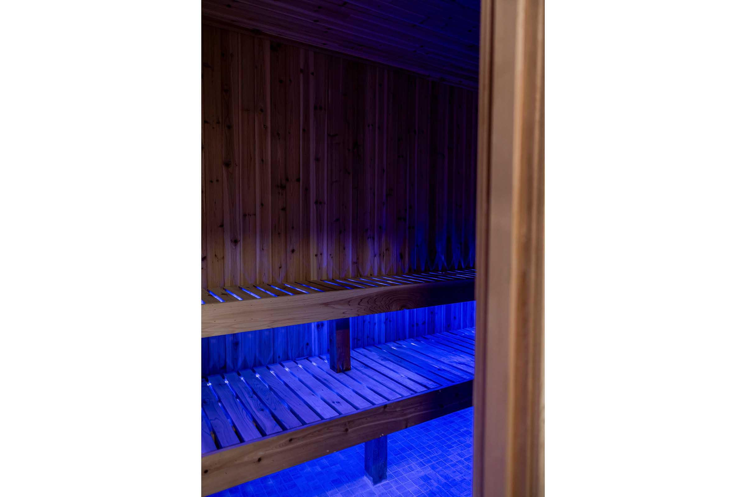 View of the interior of a sauna, featuring wooden benches and walls, illuminated by soft blue lighting. The space appears inviting and tranquil, ideal for relaxation and wellness. The scene conveys a sense of warmth and calmness, with a focus on natural materials.