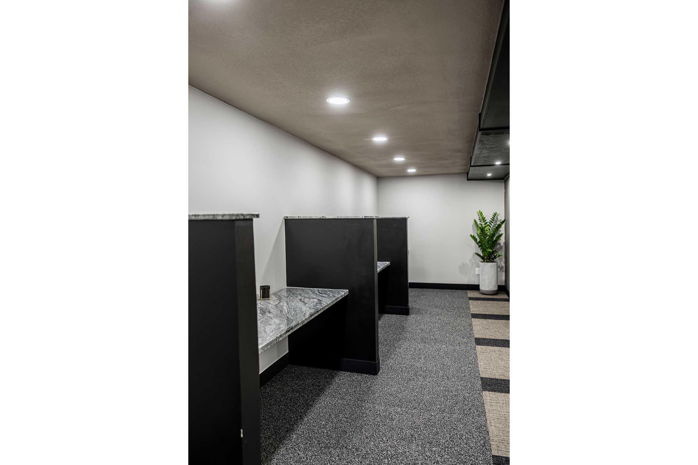 Interior view of a minimalist space featuring three dark partitioned areas with gray countertops, set against a plain white wall. The floor is covered in dark carpeting with a striped design. A potted plant is visible in the corner, and overhead lighting provides a bright atmosphere.