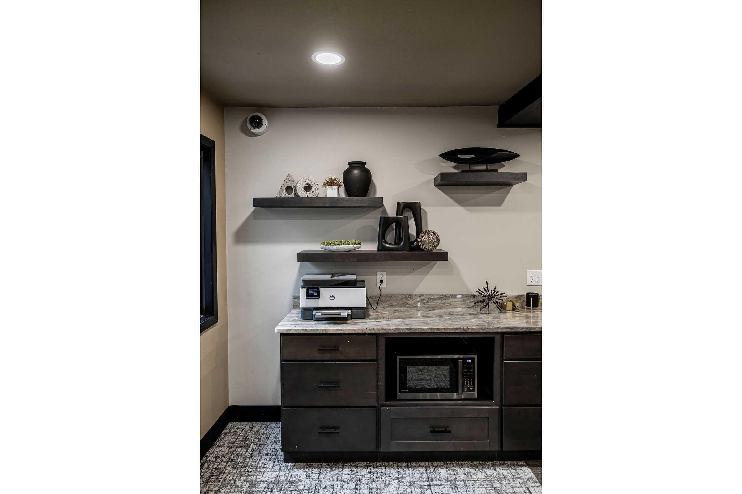 A modern kitchen scene featuring dark cabinets and a granite countertop. Open shelves above hold decorative items, including vases and art pieces. A microwave is built into the cabinetry, with muted lighting illuminating the space. The overall design is sleek and contemporary.