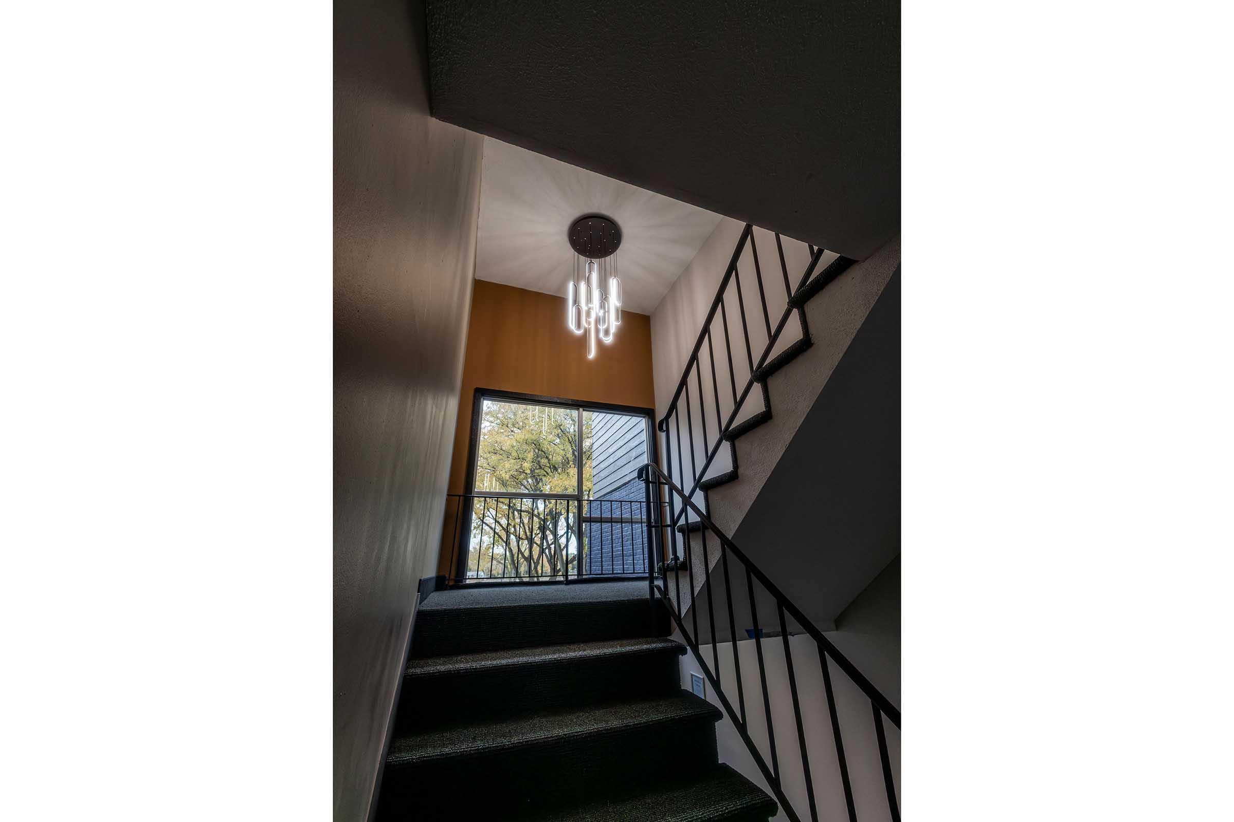 A staircase leading upward, featuring a modern chandelier hanging from the ceiling. The walls are painted in warm tones, and there is a window at the top of the stairs, allowing natural light to enter. The staircase has dark-colored steps and metal railings.