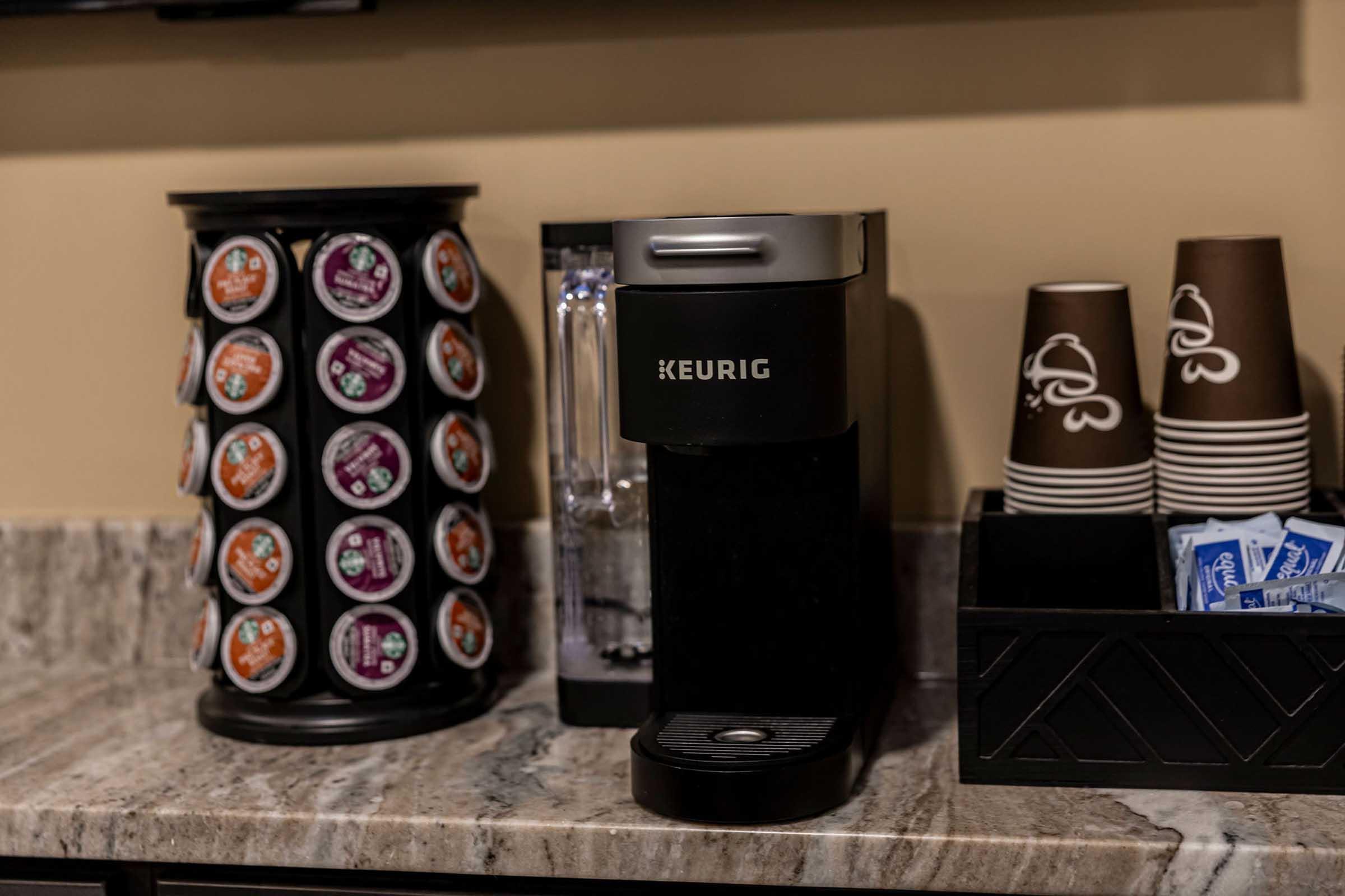 A countertop coffee station featuring a Keurig coffee maker, a carousel displaying various coffee pods, disposable coffee cups, and a container with sugar packets. The setting has a modern design with a marble surface and neutral-colored walls.