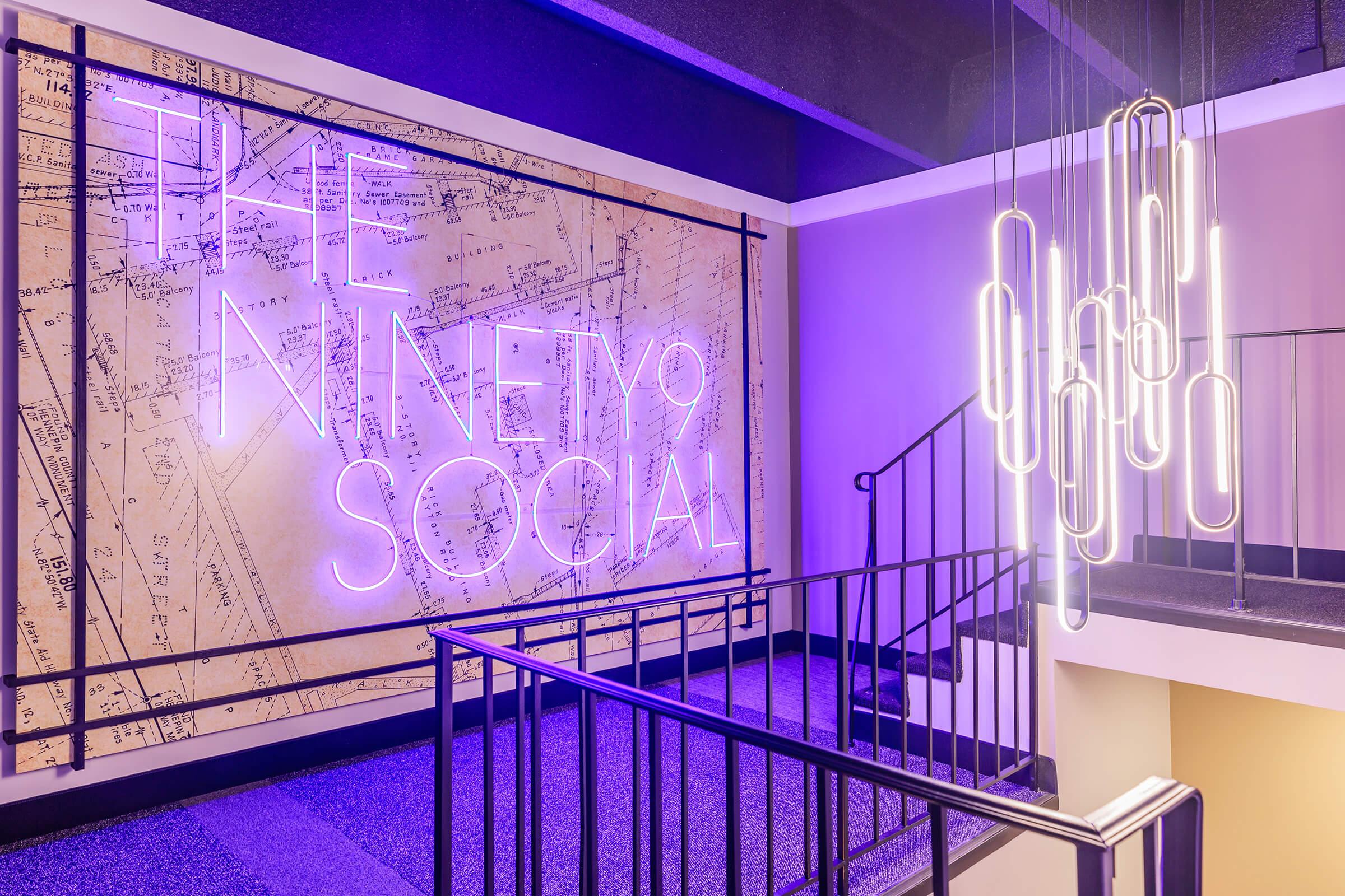 A modern interior view featuring a staircase with a stylish black railing. The wall is adorned with a large illuminated sign that reads "THE NINETY SOCIAL" in neon letters against a textured backdrop, and there are unique, contemporary light fixtures hanging from the ceiling, adding to the trendy atmosphere.