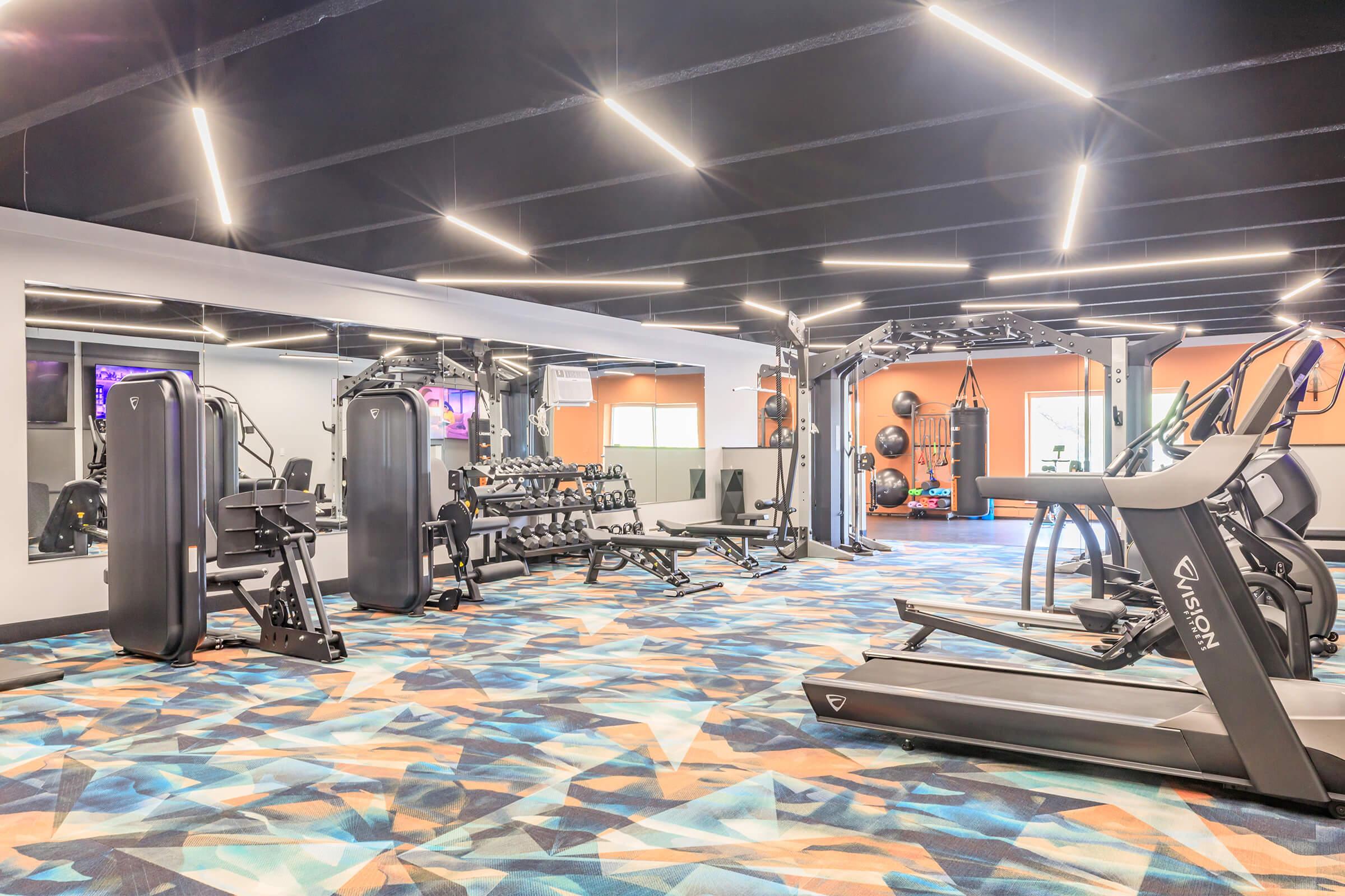 A modern gym featuring various exercise equipment, including treadmills, weight machines, a free weights area, and a cable machine. The space has vibrant flooring and bright lighting, creating an inviting workout environment. Large mirrors reflect the area, enhancing the sense of openness.