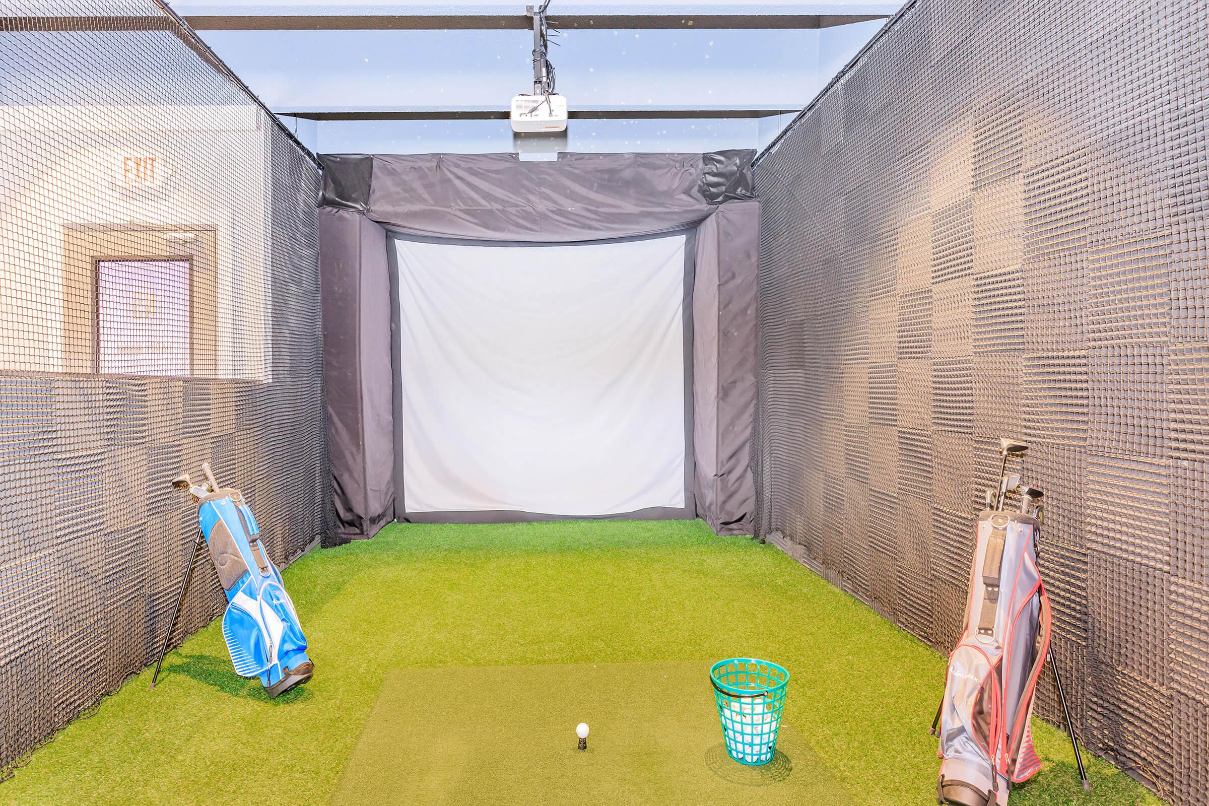 Indoor golf practice area featuring artificial turf, a projector screen at the front, a golf ball positioned on a tee, and two golf bags with clubs leaning against the walls. The space is enclosed with netting for safety and has a clean, organized appearance.