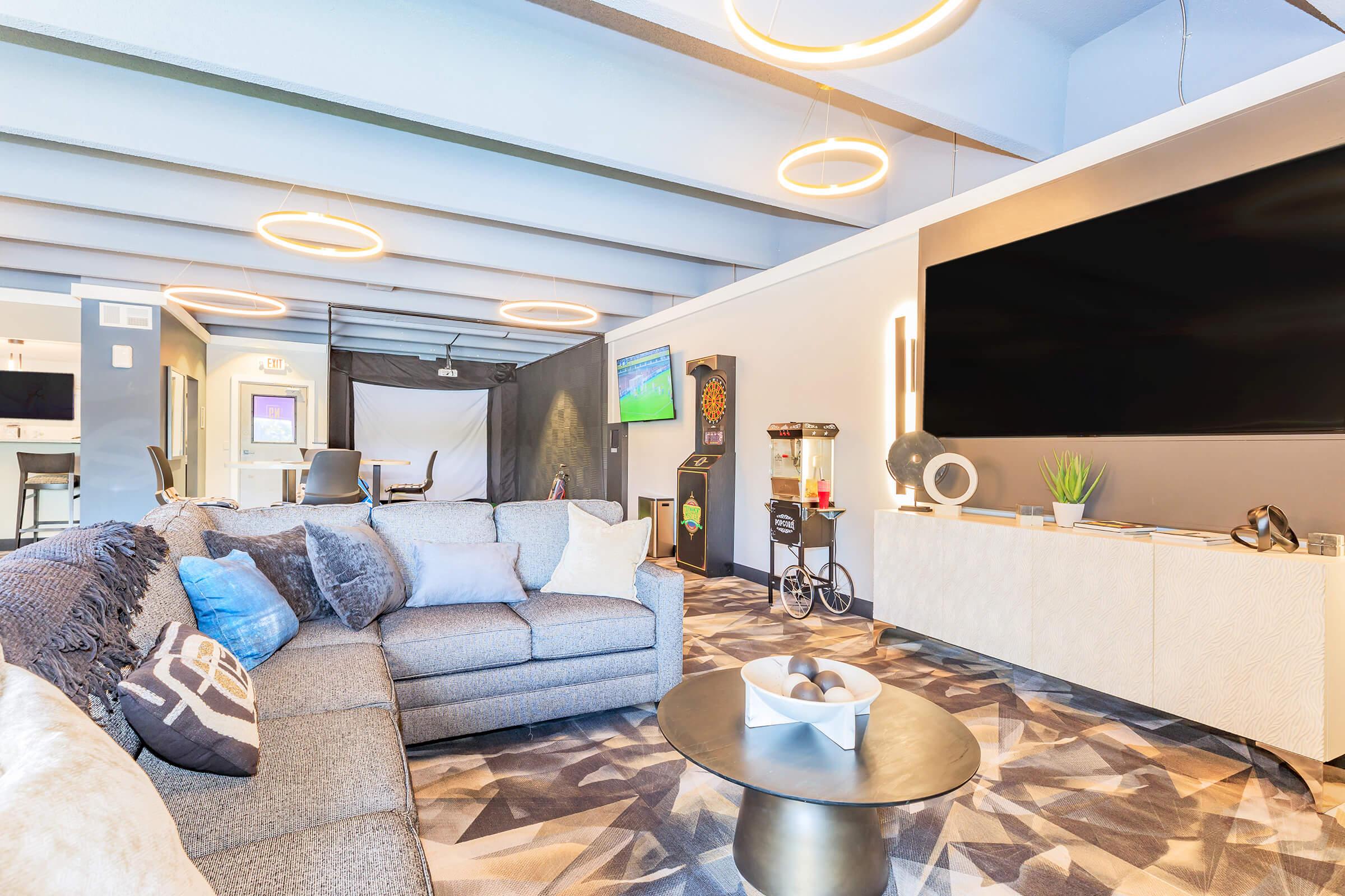 A modern living room featuring a large gray sofa adorned with decorative pillows, a stylish coffee table, and a flat-screen TV mounted on a light-colored cabinet. Circular pendant lights hang from the ceiling, and there are accent pieces like a plant and a dartboard in the background, creating a cozy atmosphere.