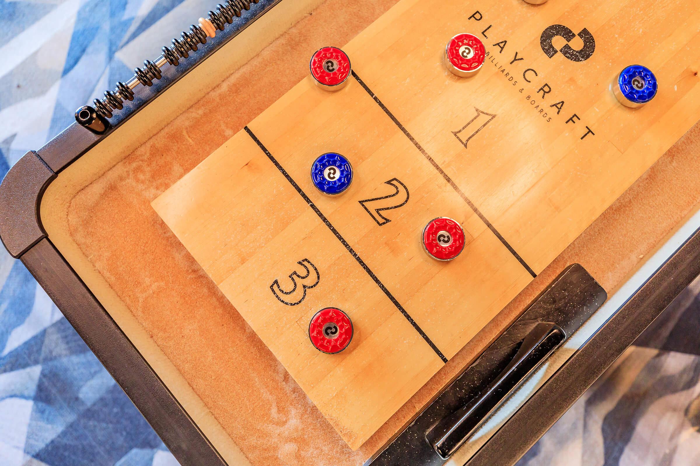A close-up view of a Playcraft shuffleboard table featuring a wooden playing surface with numbered sections. Red and blue pucks are positioned on the board, and the edges of the table show a sleek design with a dark frame. The background has a patterned fabric.