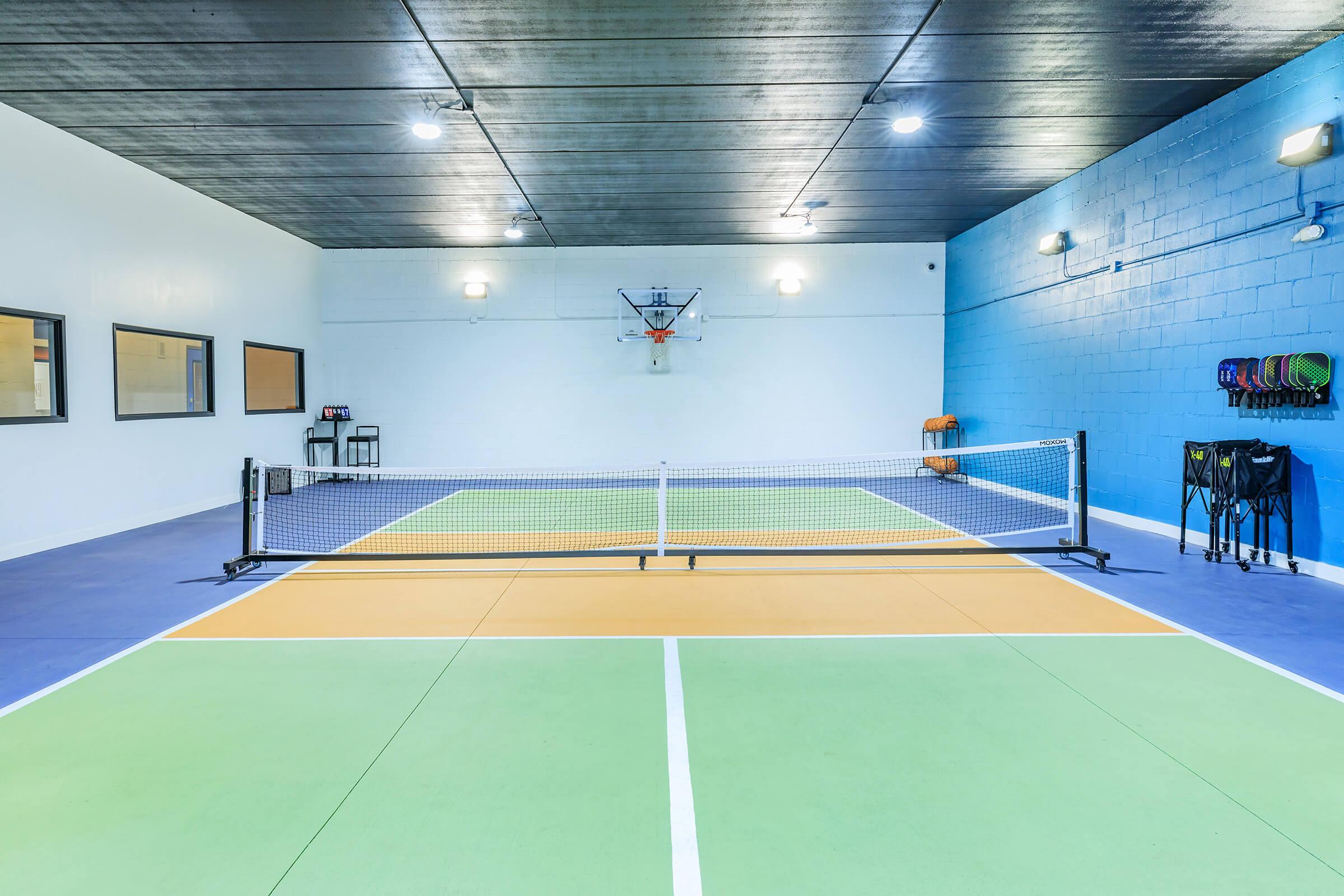 Indoor sports facility featuring a multi-sport court with a basketball hoop and a netted area. The court is divided into green, orange, and blue sections, with walls painted in white and blue. There are benches and equipment storage visible in the background. Bright overhead lights illuminate the space.