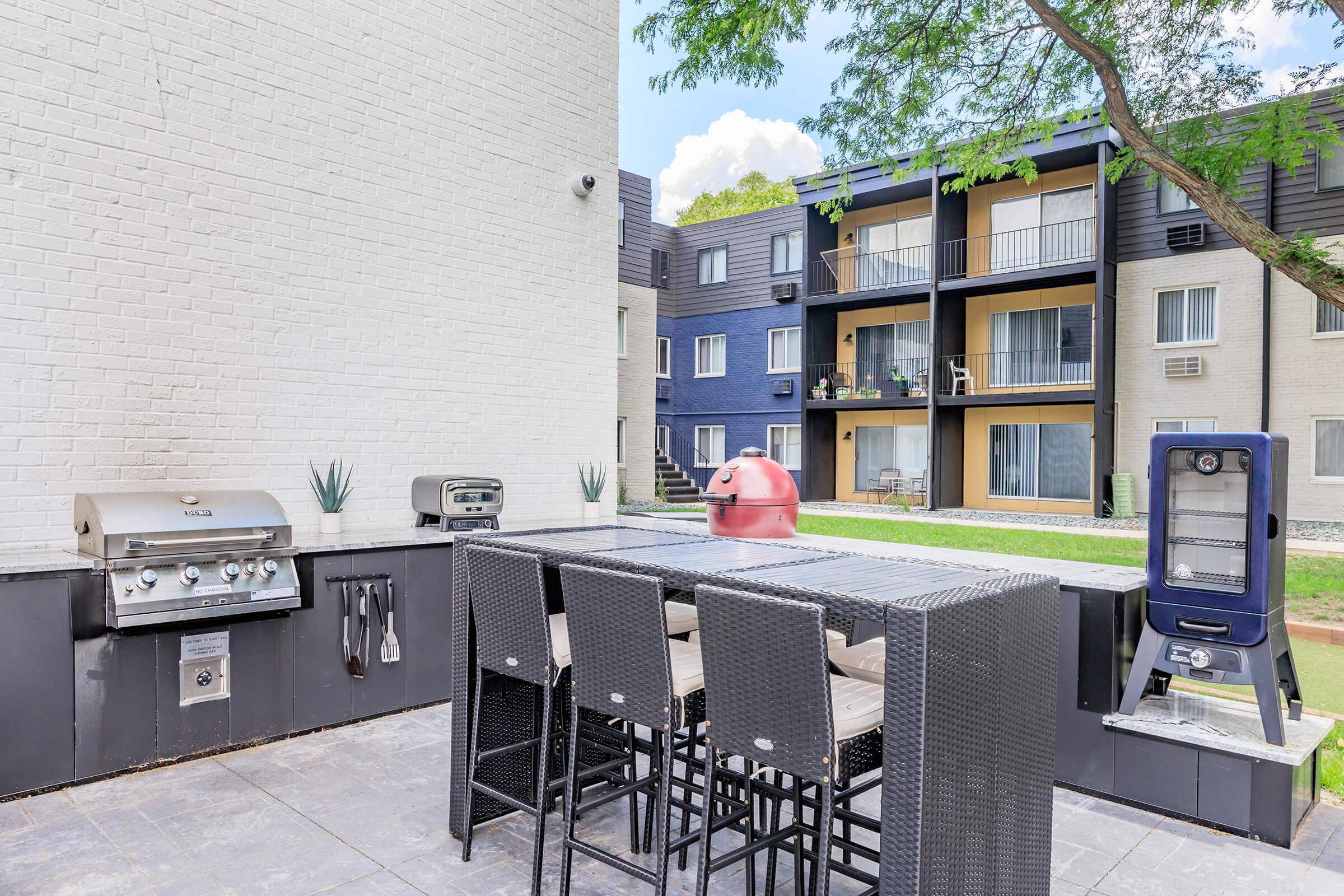 An outdoor patio area featuring a modern grill setup, a large dining table with black wicker chairs, and a bright red smoker. In the background, there are several apartment units with balconies and green lawn space. Trees provide shade, creating a comfortable outdoor dining environment.