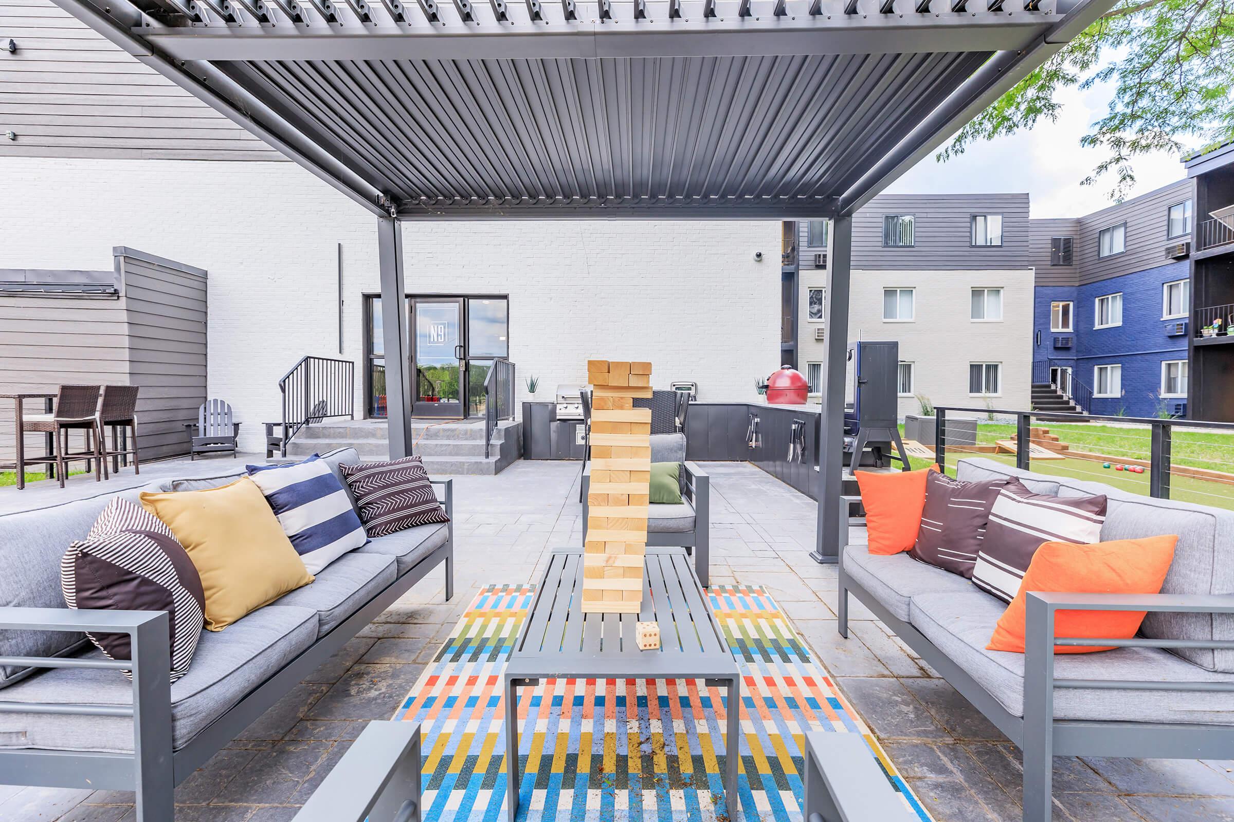 Outdoor lounge area featuring gray sofas with colorful cushions, a table with a Jenga game stacked in the center, and a vibrant rug. In the background, there are outdoor grills and modern apartment buildings, creating a relaxed and inviting atmosphere for social gatherings.