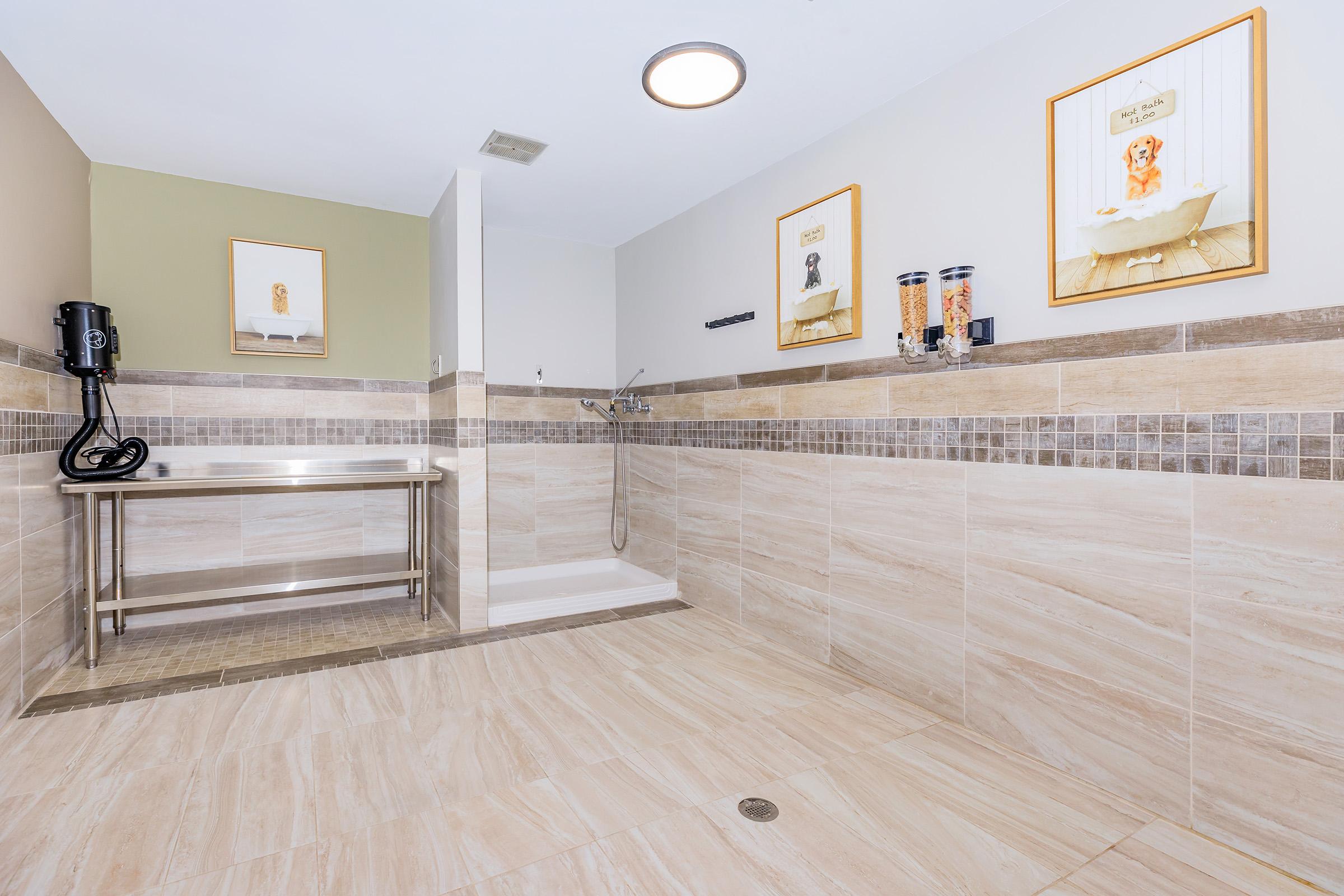 A modern and spacious pet grooming area featuring light-colored tiled walls and floor. There is a grooming table in the center, a shower area with a handheld shower head, and complimentary grooming supplies displayed on the wall. Two framed pet-themed artworks are hung on the walls, enhancing the inviting atmosphere.