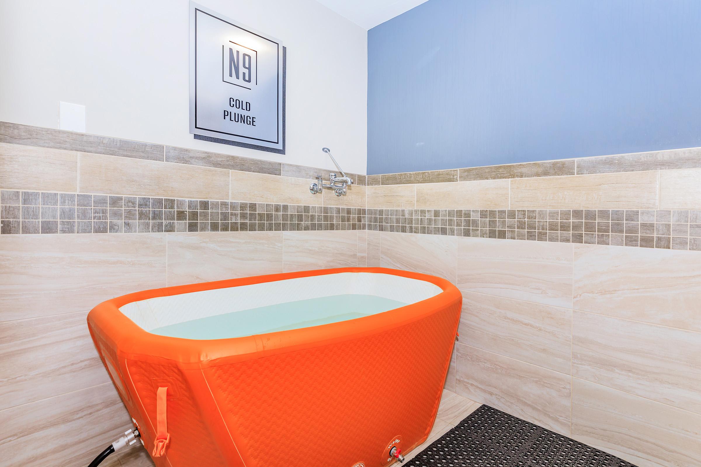 A vibrant orange cold plunge tub is situated in a tiled room. The tub is filled with water and is accompanied by a wall-mounted faucet. Above it, there is a framed sign reading "N9 COLD PLUNGE." The walls are a mix of light and dark tiles, creating a modern and relaxing atmosphere.