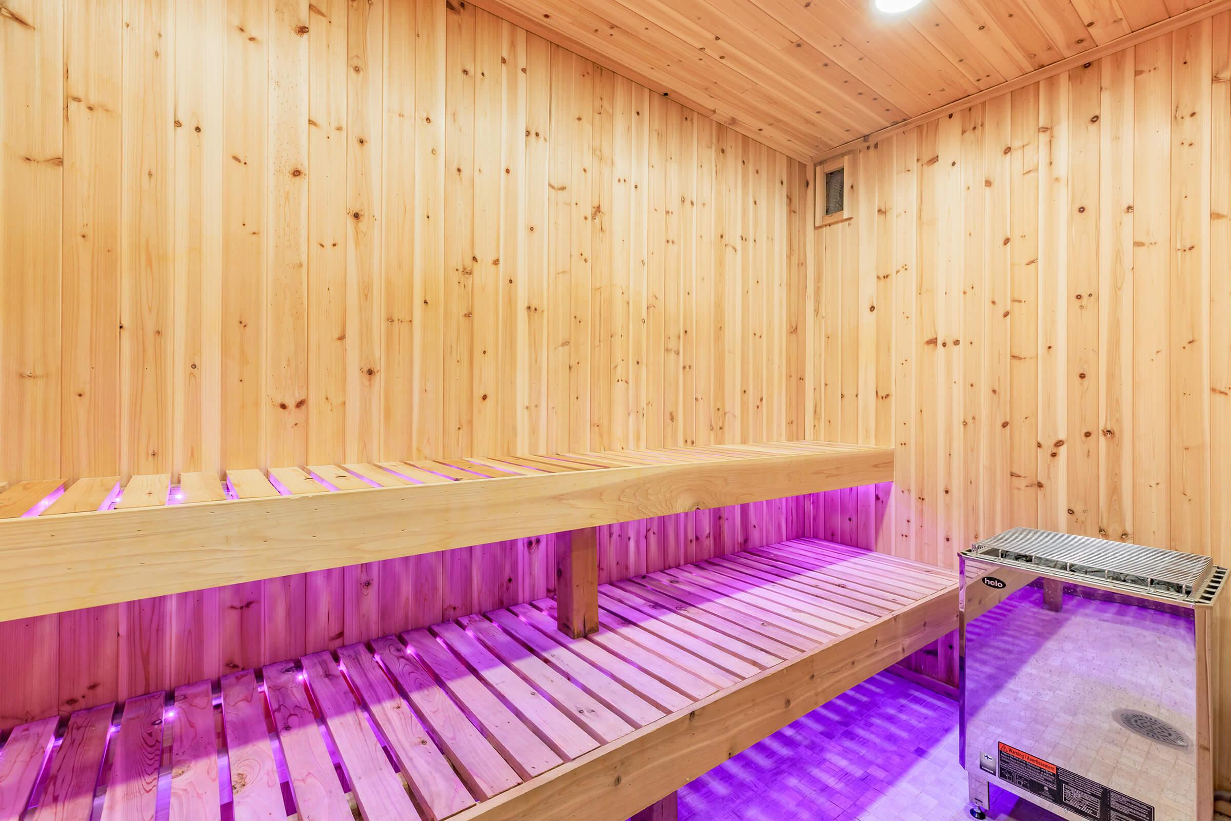 A wooden sauna interior featuring two benches, one above the other, illuminated with purple lighting. The walls are lined with wooden planks, creating a warm, cozy atmosphere. A sauna heater is positioned on the right side of the image, completing the serene setting.