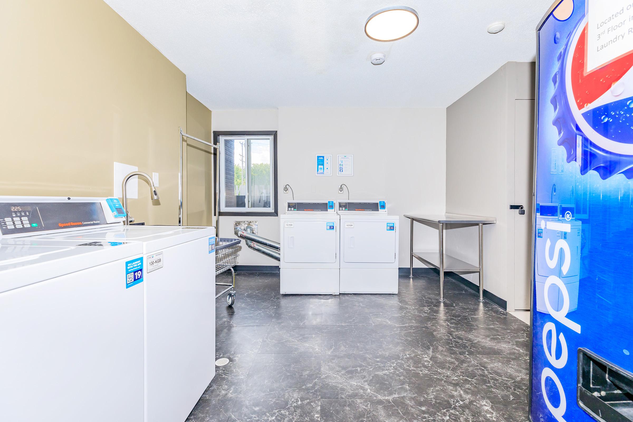 A clean and modern laundry room featuring two white washing machines and dryers, a stainless steel table, and a utility sink. The floor is dark and glossy, with a window providing natural light. A vending machine for laundry supplies is visible on the right.