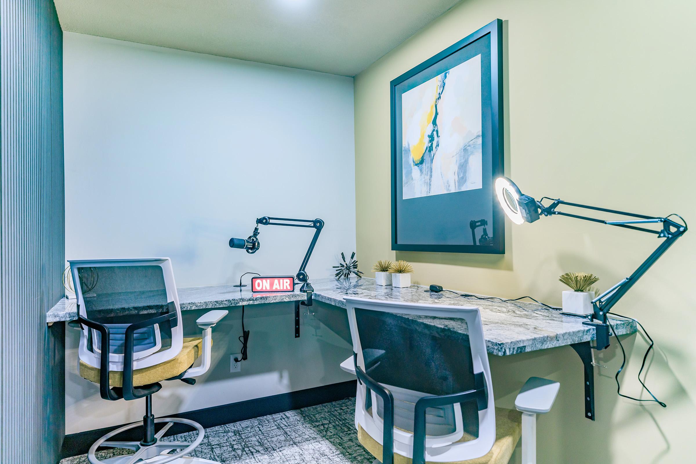 A modern workspace featuring two ergonomic chairs and a marble-top desk. The desk has adjustable arms with studio lights and a microphone. A small decorative plant is on the desk, and a framed abstract artwork hangs on the wall. A sign reading "ON AIR" is illuminated on the desk. Soft lighting enhances the inviting atmosphere.