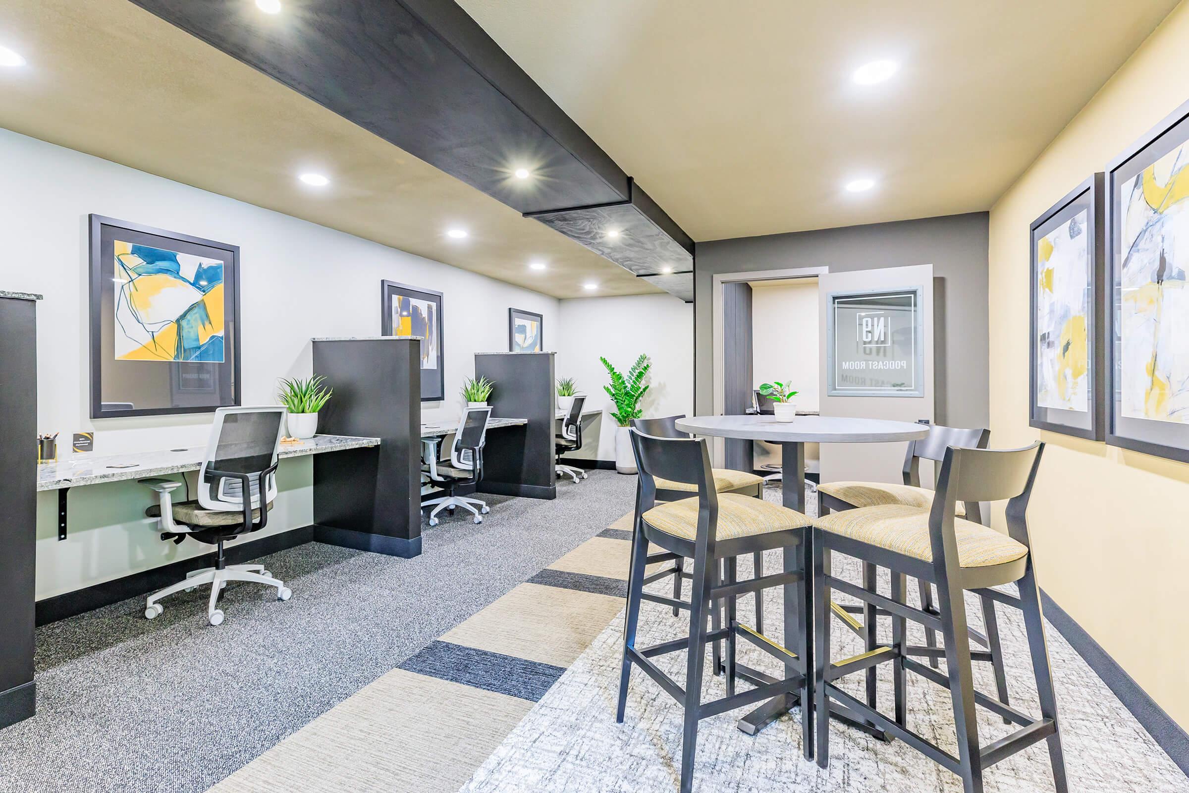 A modern, well-lit office space featuring several workstations with ergonomic chairs. There are decorative plants and artwork on the walls, and a circular table with high chairs in the center, creating a collaborative environment. The flooring combines carpet and a patterned area rug.