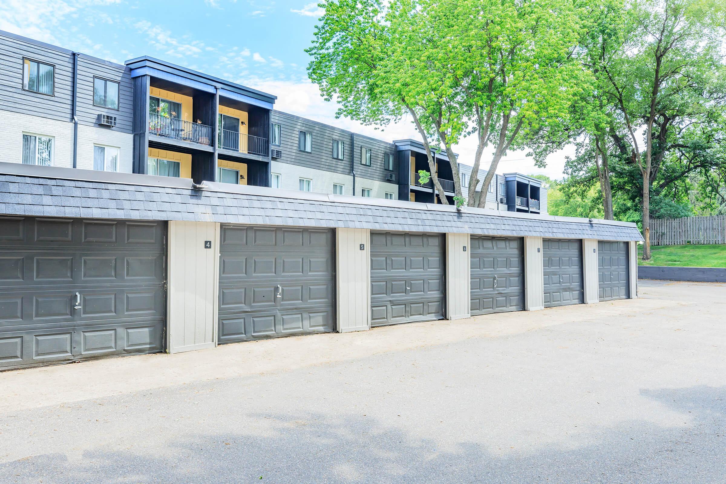 A row of closed garage doors in front of a two-story apartment building. The building features a modern design with balconies and large windows, surrounded by green trees and open sky. The pavement in front of the garages is clean and well maintained.