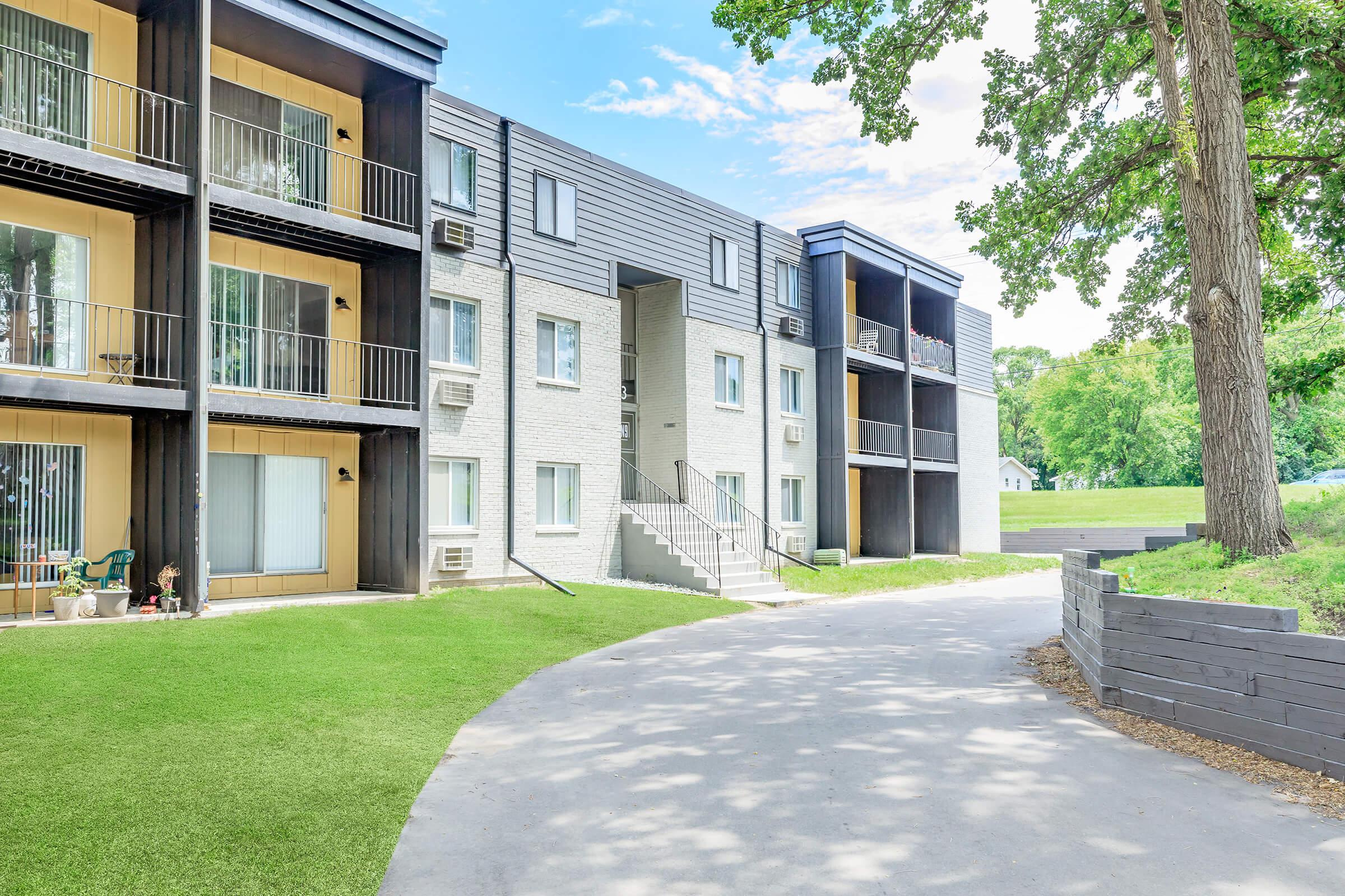 A modern multi-story apartment building featuring various balconies, large windows, and a landscaped pathway. The building is surrounded by greenery and trees, with a blue sky and scattered clouds in the background. The setting appears peaceful and residential.