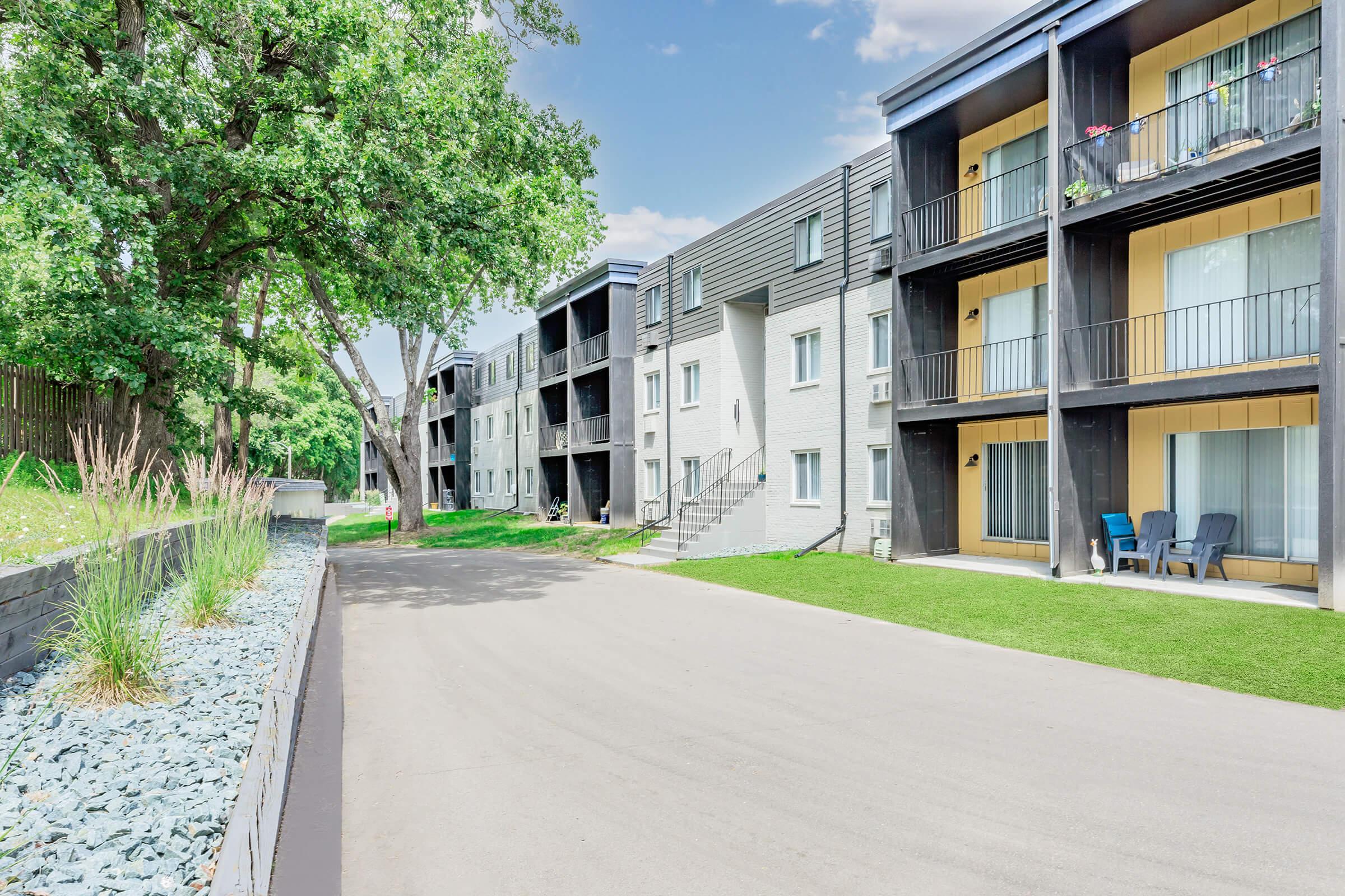A view of a modern apartment complex featuring three-story buildings with balconies. The walkway is lined with greenery and decorative rocks, and there are trees on one side. The sky is partly cloudy, creating a bright and inviting atmosphere.